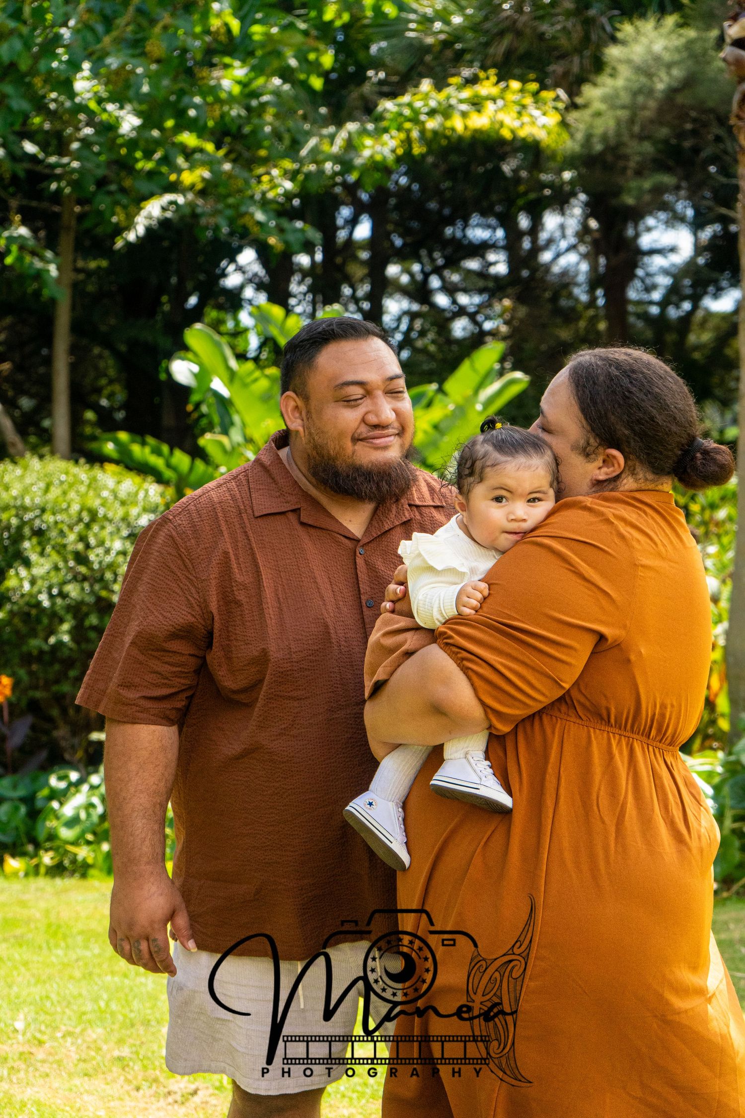 Loving Pacific Island family with father holding baby girl while mother kisses child in garden.