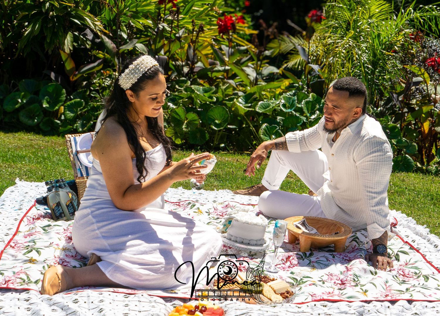 Couple in white outfits enjoying a romantic outdoor picnic on a floral blanket in a lush green garden.