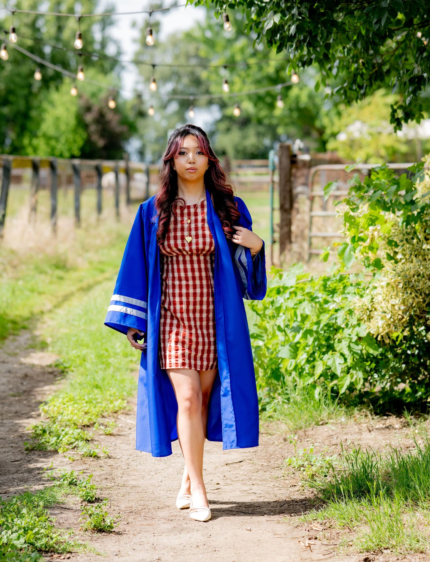 Young woman walks a garden path in a blue graduation gown over a red gingham dress with string lights above.