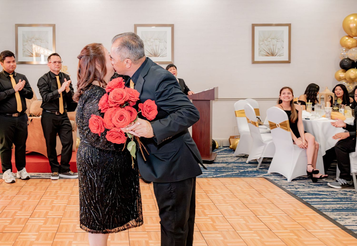 Couple shares romantic kiss with red roses at elegant black and gold 60th birthday party celebration.