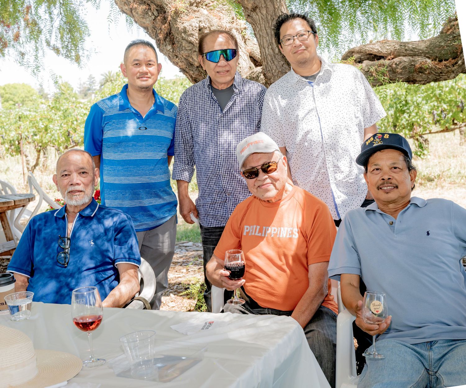Six men posing together outdoors under a tree at a sunny gathering, some holding wine glasses at a table.