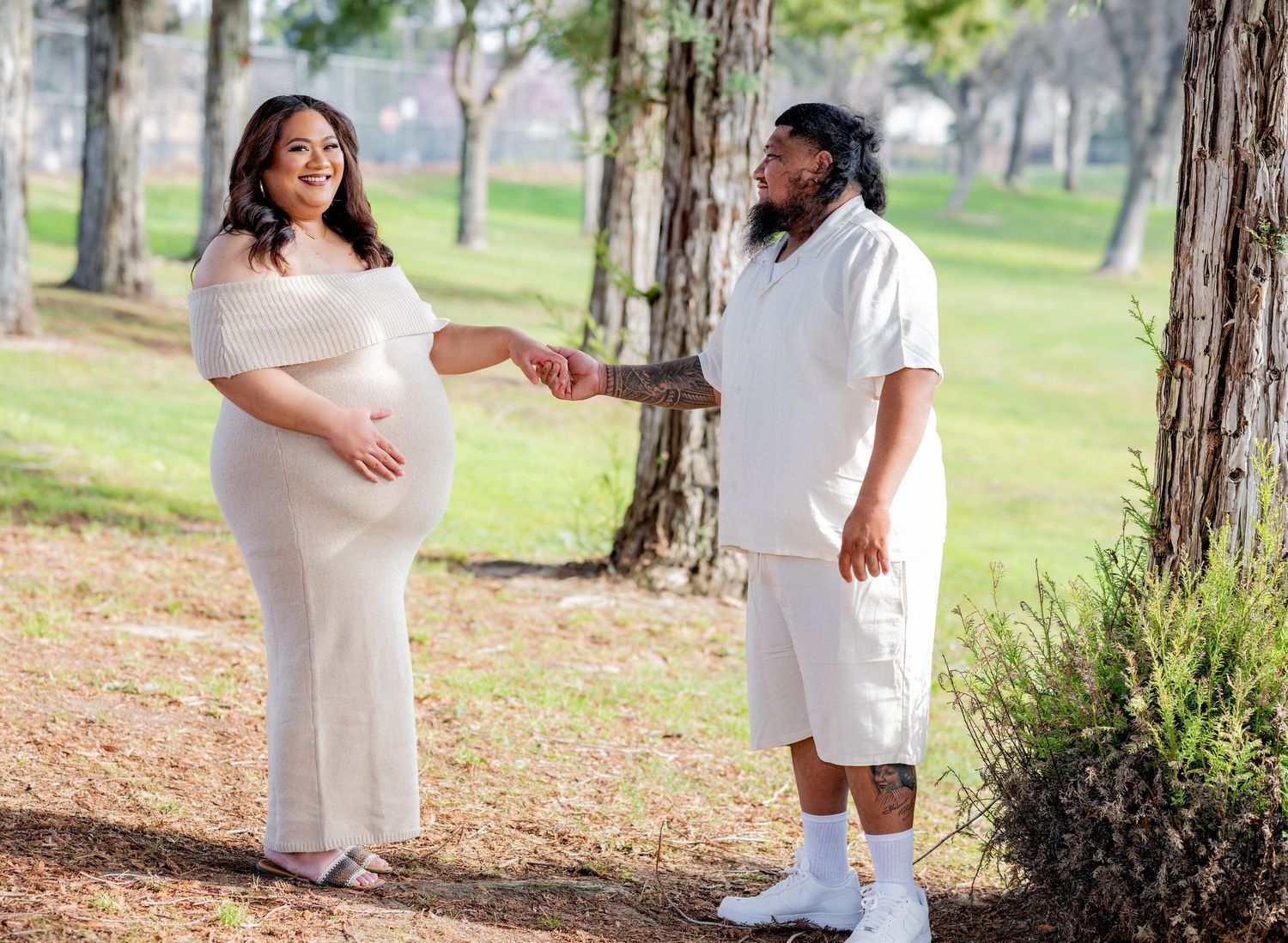 Pregnant woman in cream dress holds hands with bearded partner in white outfit among park trees.