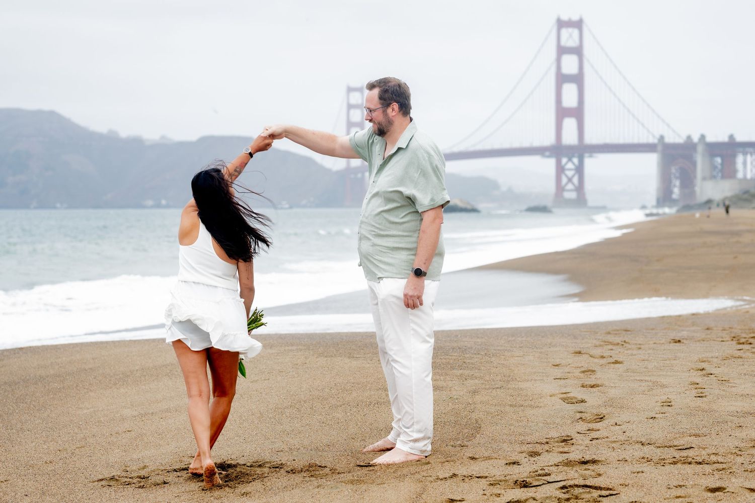 Couple sharing a dance on Baker Beach shoreline with the Golden Gate Bridge framed behind them.