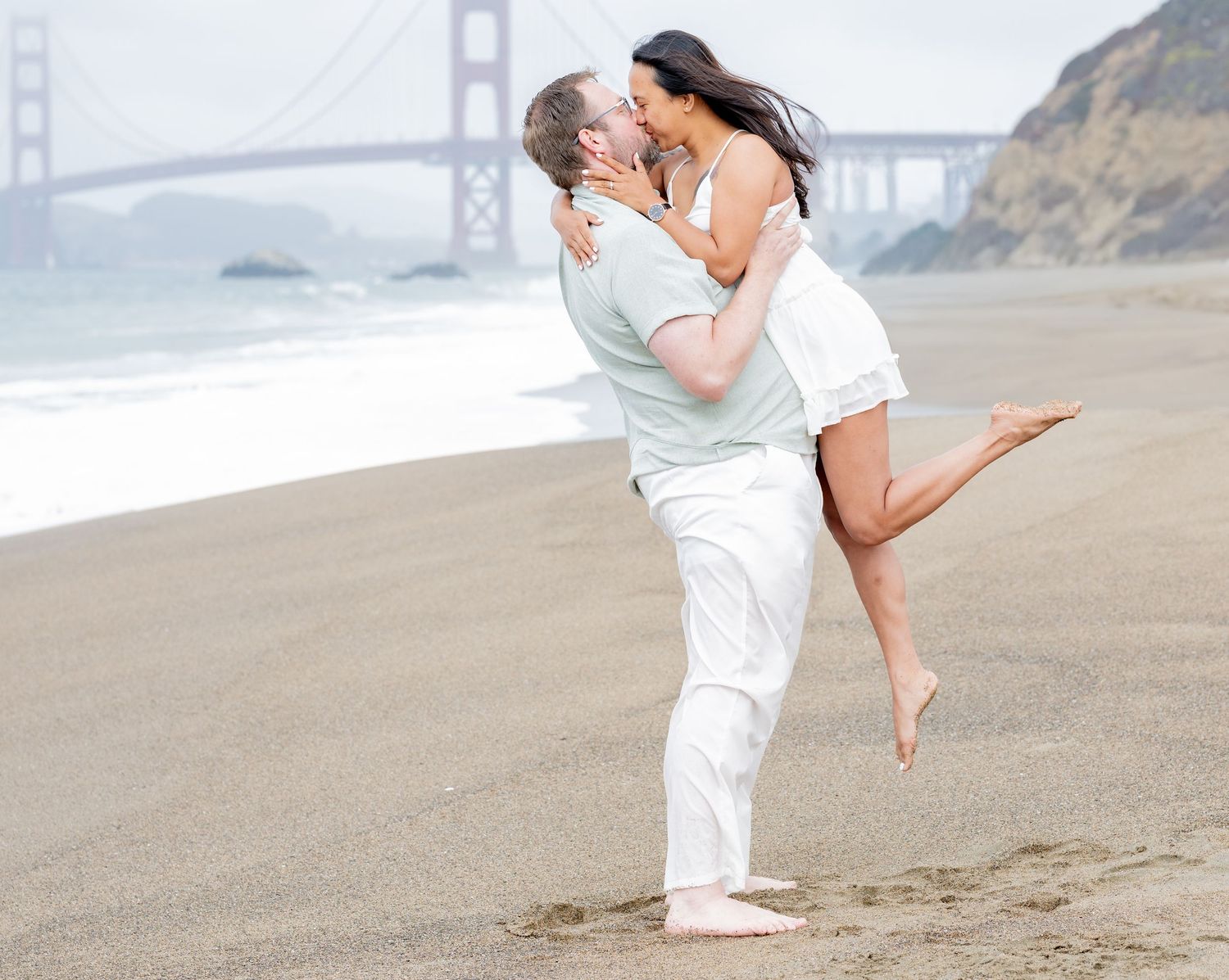 Romantic couple shares a kiss on Baker Beach with the Golden Gate Bridge visible in the background.