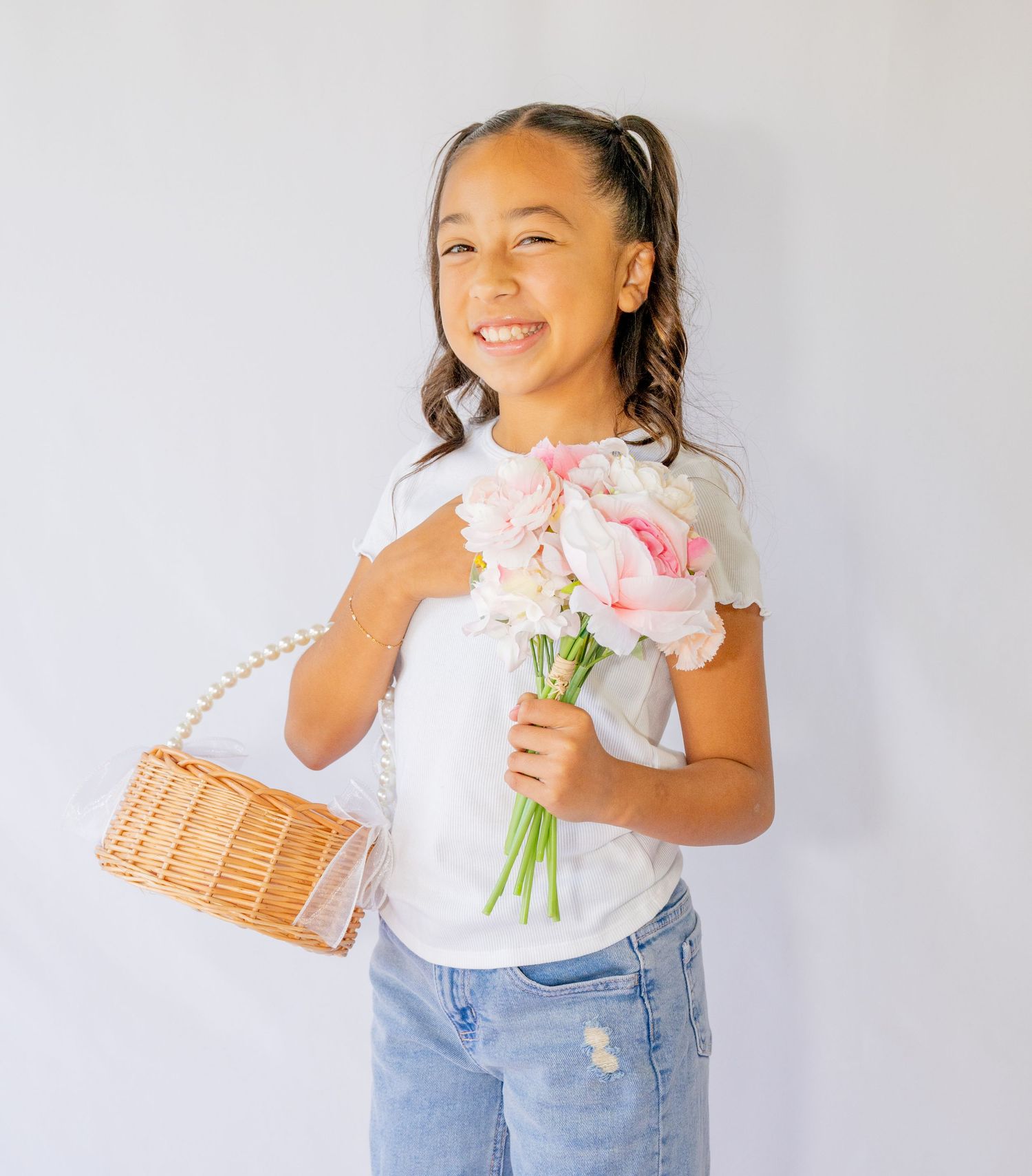 Smiling girl holding pink flower bouquet and wicker purse for birthday portrait on white background.