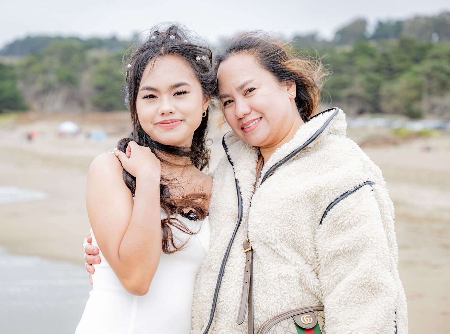 Two women smile closely together for a portrait on a sandy beach near the water.
