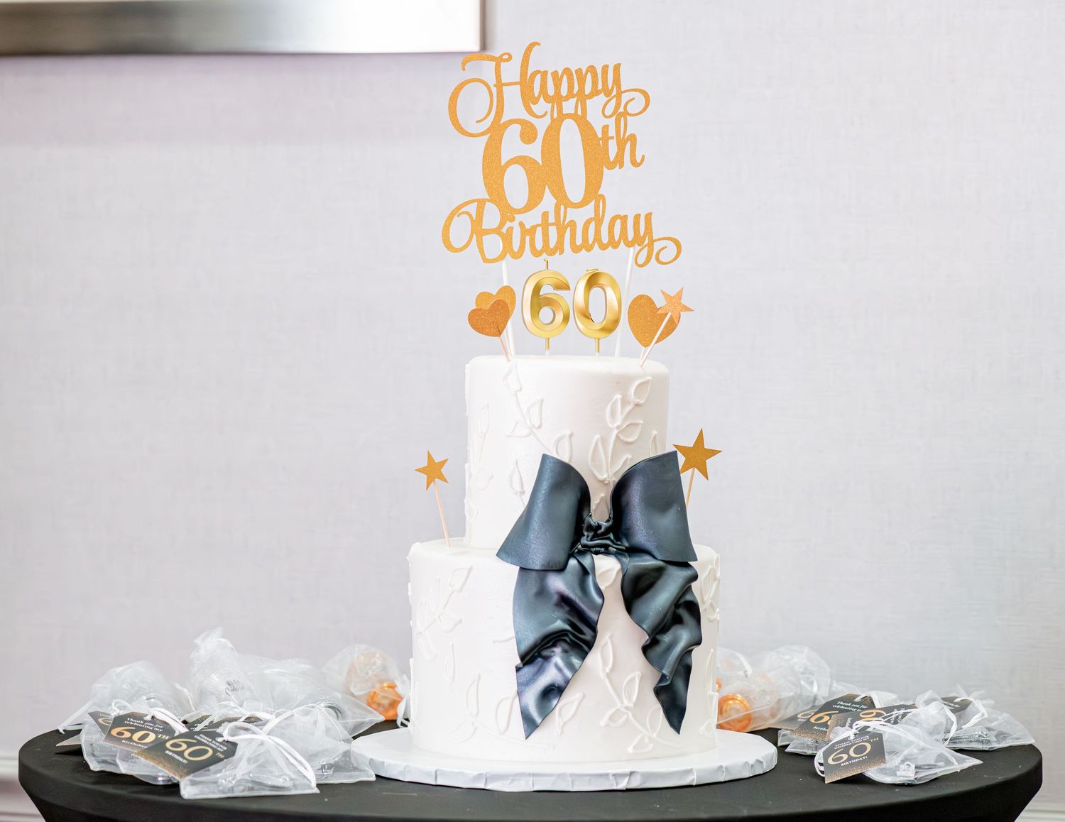 Elegant two-tier white birthday cake with gold Happy 60th Birthday topper and navy blue bow.