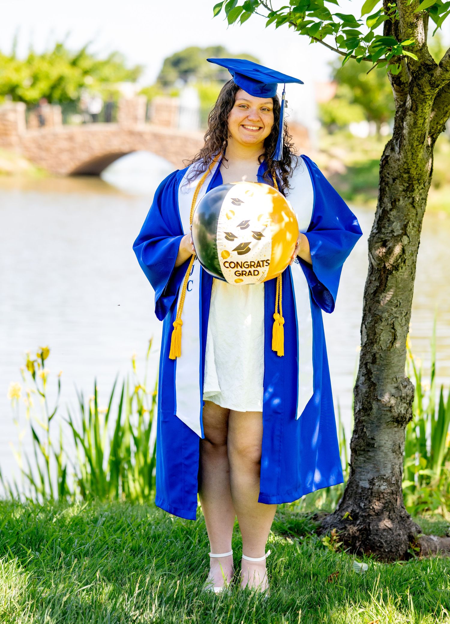 Graduate in blue cap and gown holds Congrats Grad balloon near a scenic pond.