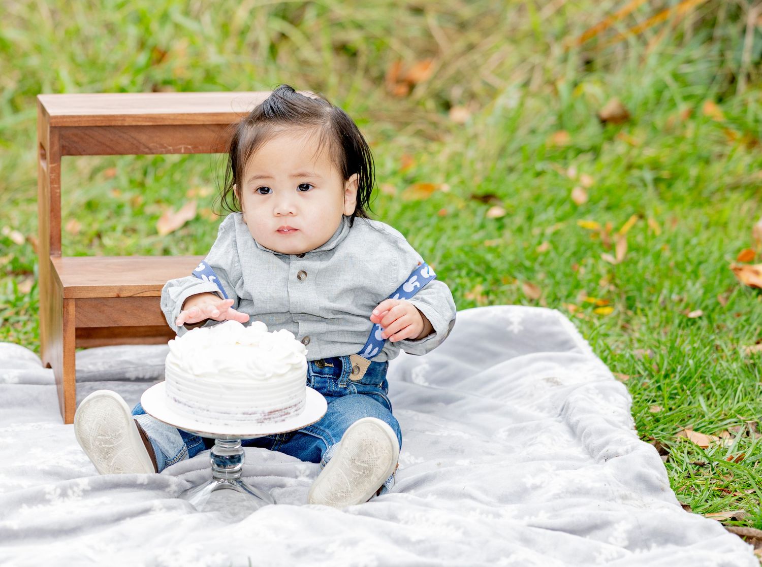 Baby boy in denim and suspenders reaches toward white smash cake on glass stand outdoors.