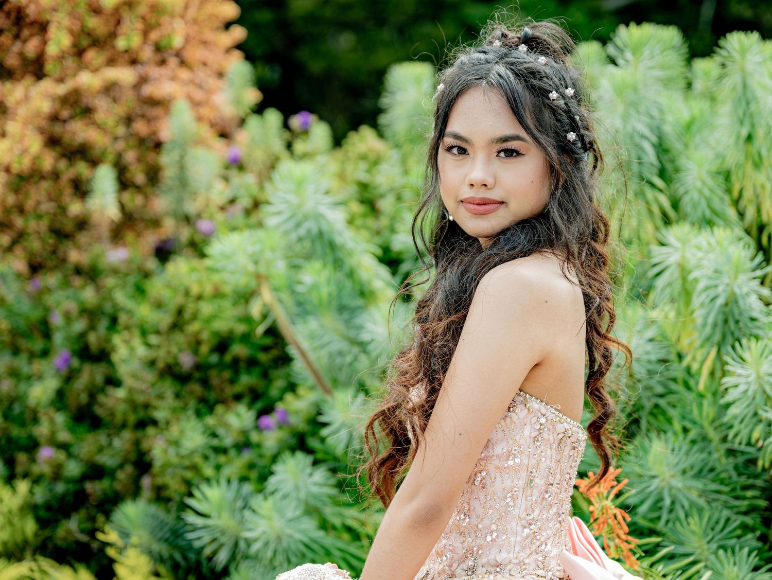 Teen girl in strapless blush quinceañera gown posing gracefully among lush garden greenery.