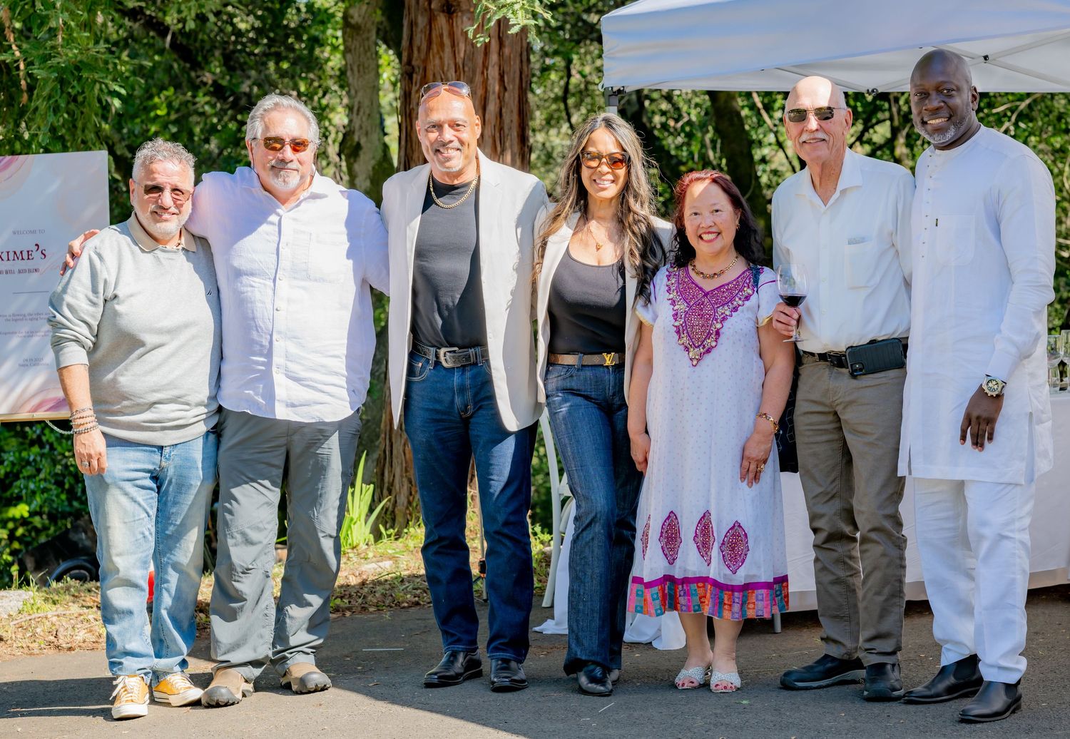 Group of seven diverse adults posing together outdoors at a sunny garden celebration event.