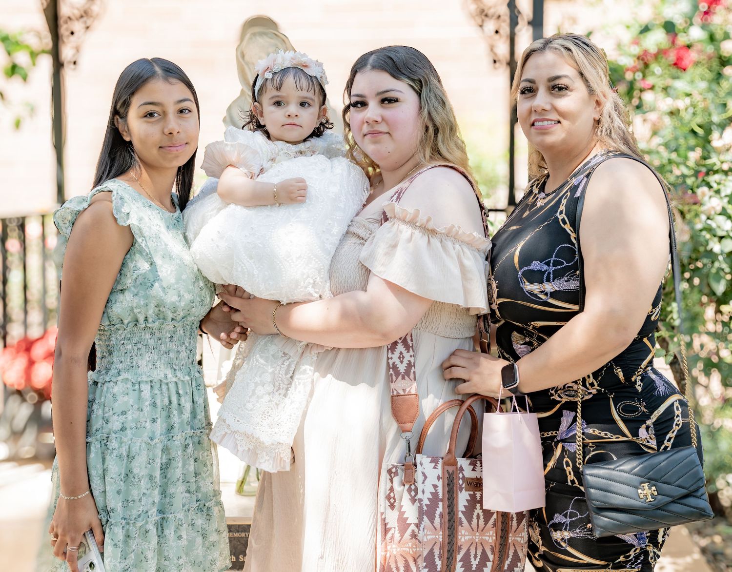 Three women and a girl pose outdoors with baby dressed in white baptism gown.
