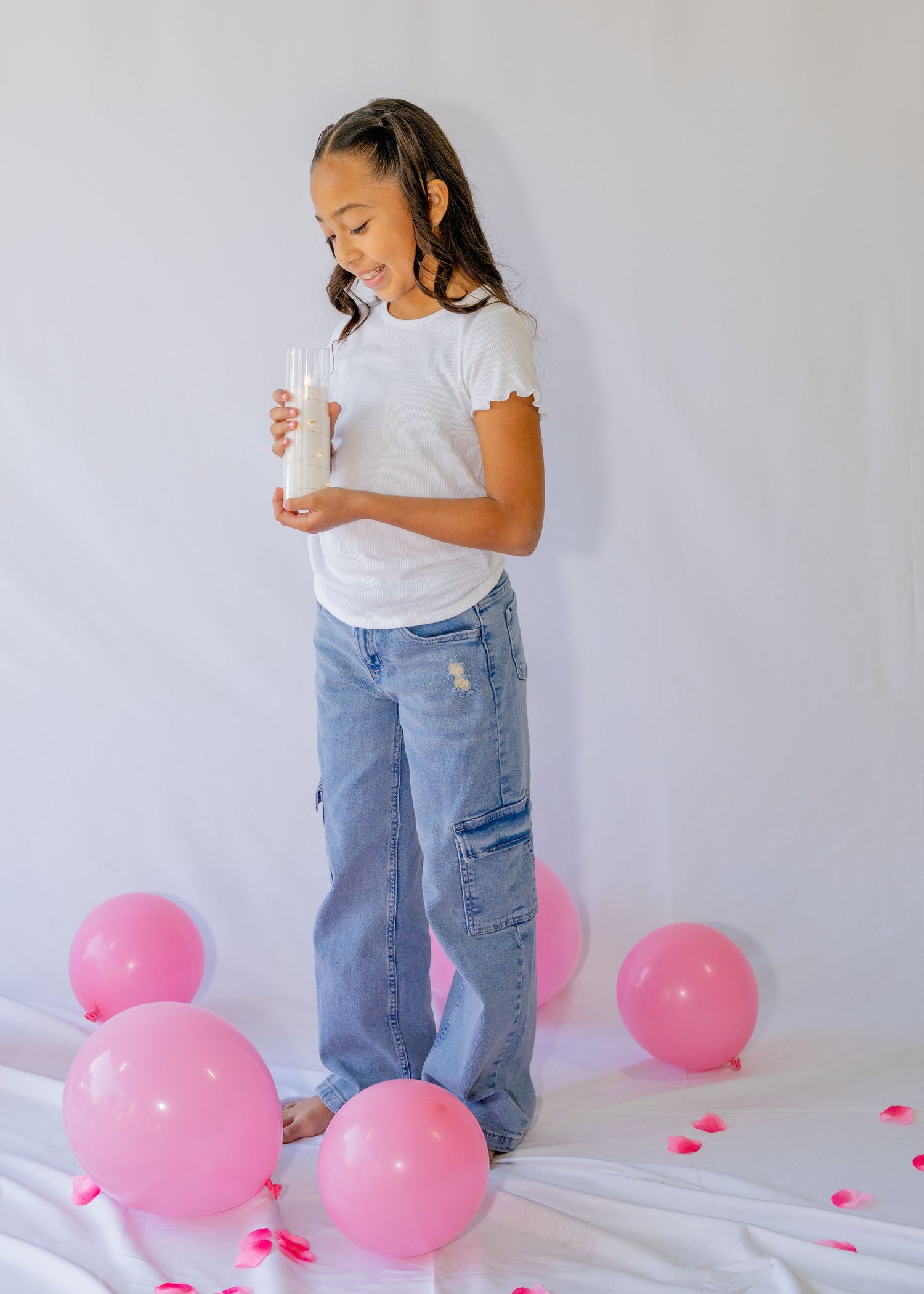 Girl in white tee and cargo jeans holds a glass amid pink balloons on white backdrop.