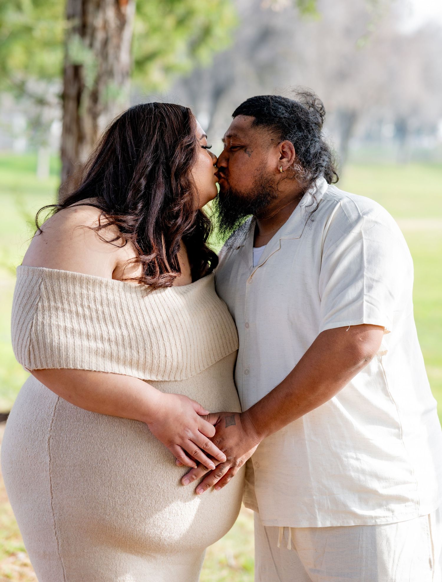 Expecting couple shares a romantic kiss in a park, with man gently holding pregnant partner's belly.