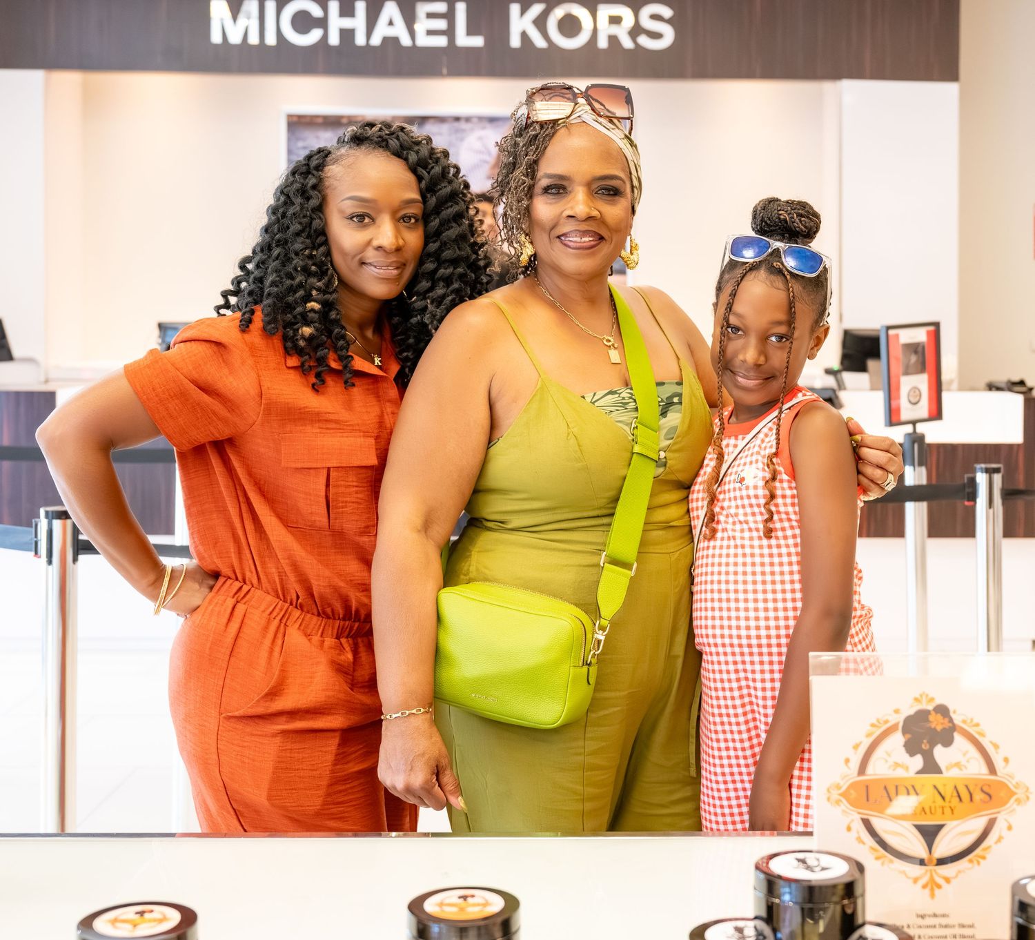 Three women smiling together in front of a Michael Kors store display, wearing colorful outfits in orange, green, and red.