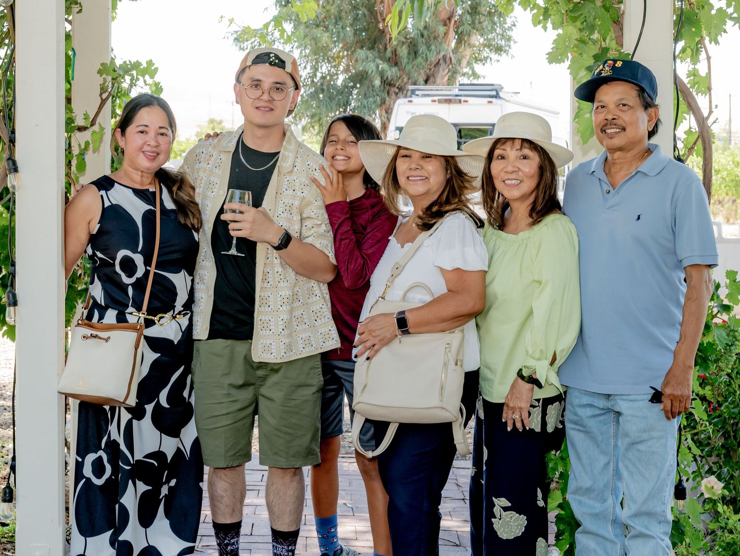 Group of seven friends posing together outdoors under a pergola at a sunny wine tasting event.