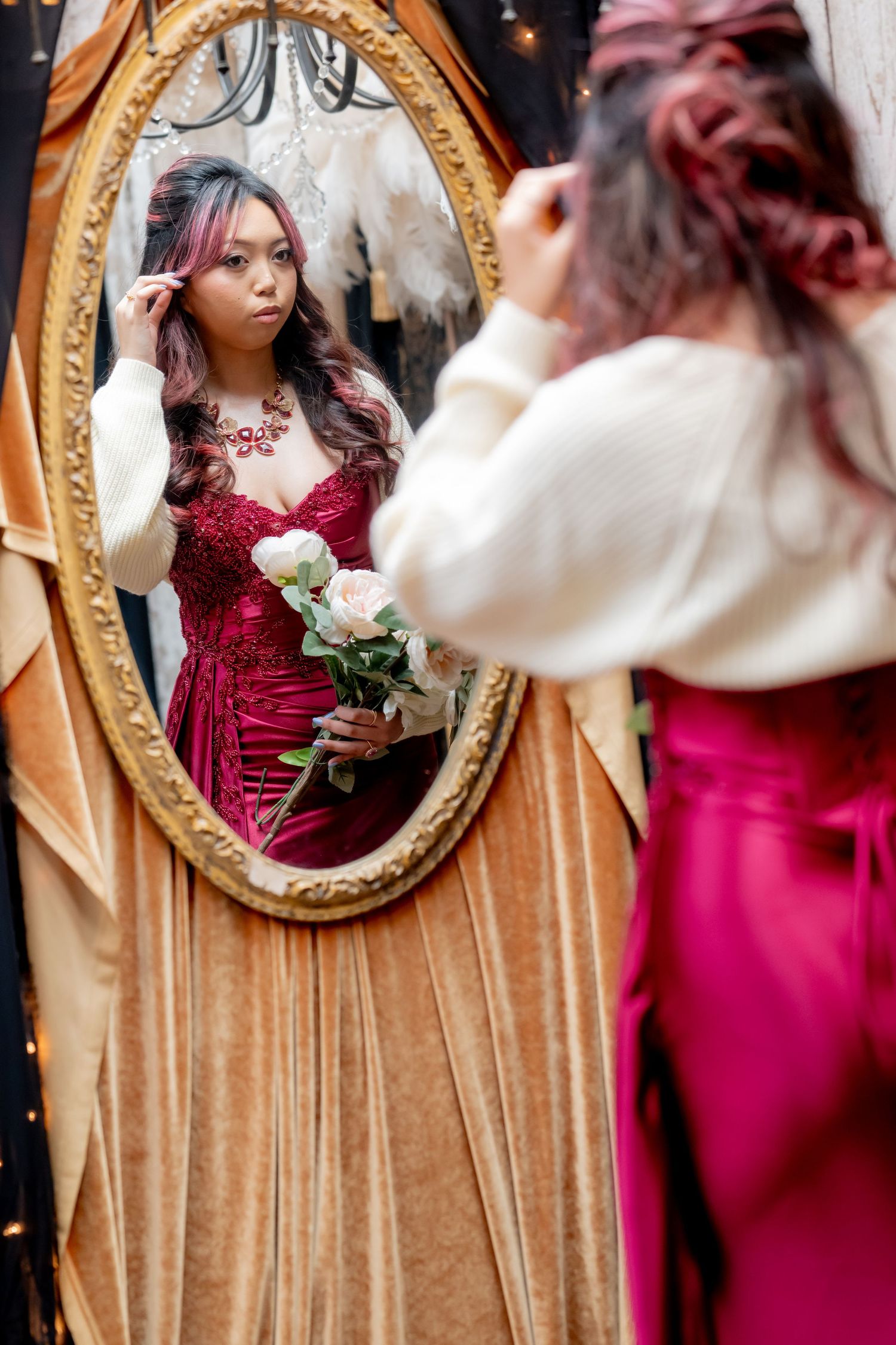 Young woman in red gown and white cardigan holding roses while checking hair in ornate oval mirror.