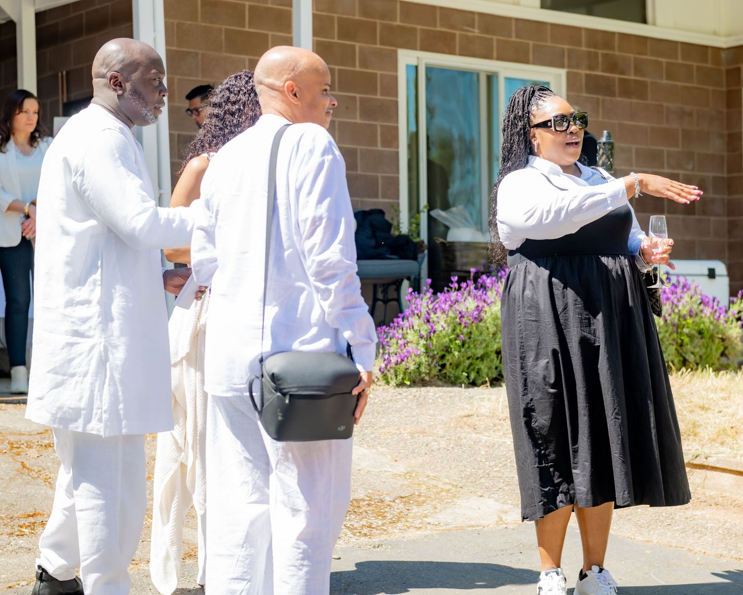 Three guests socializing outdoors at a garden event, woman in black dress gesturing while speaking to two men.