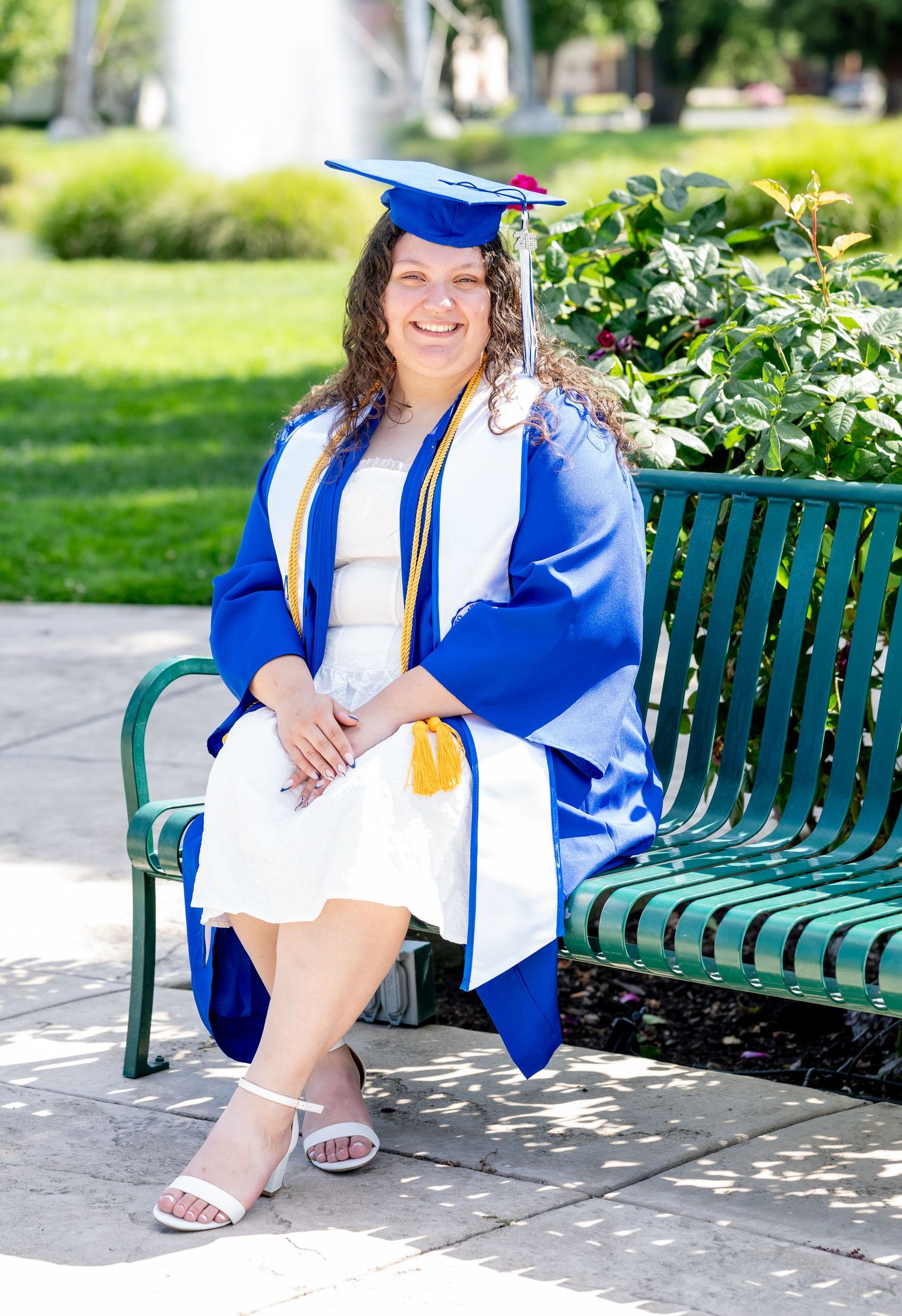 Smiling female graduate in blue cap and gown sitting on green bench in sunny park.