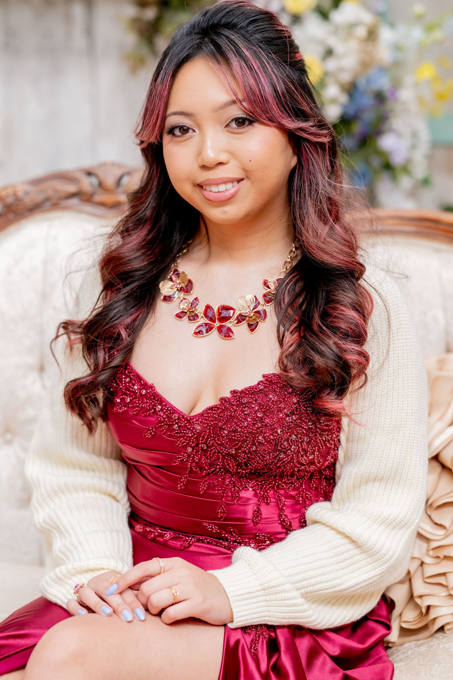 Smiling young woman in red lace gown and white cardigan wearing floral necklace on vintage sofa.