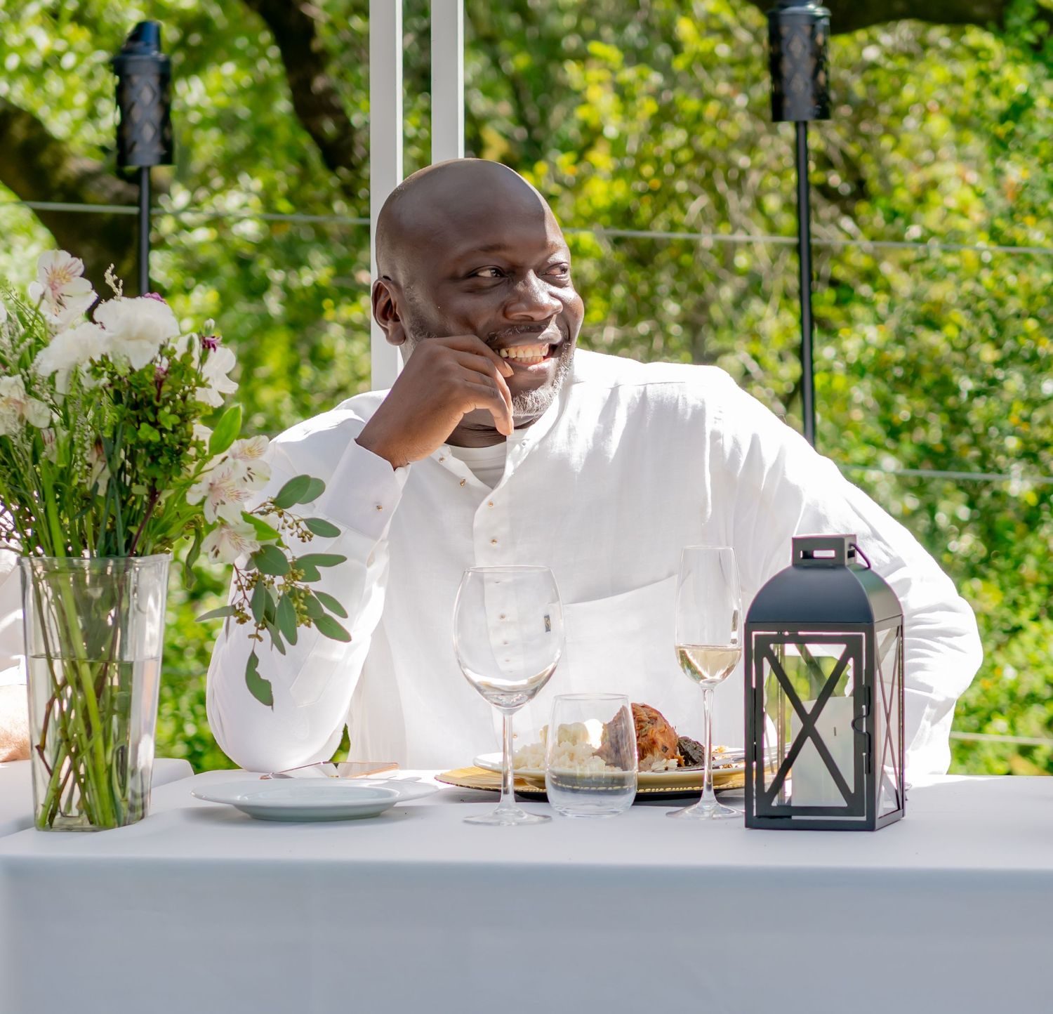 Happy man in white shirt laughing at outdoor dining table with wine glass and lantern.