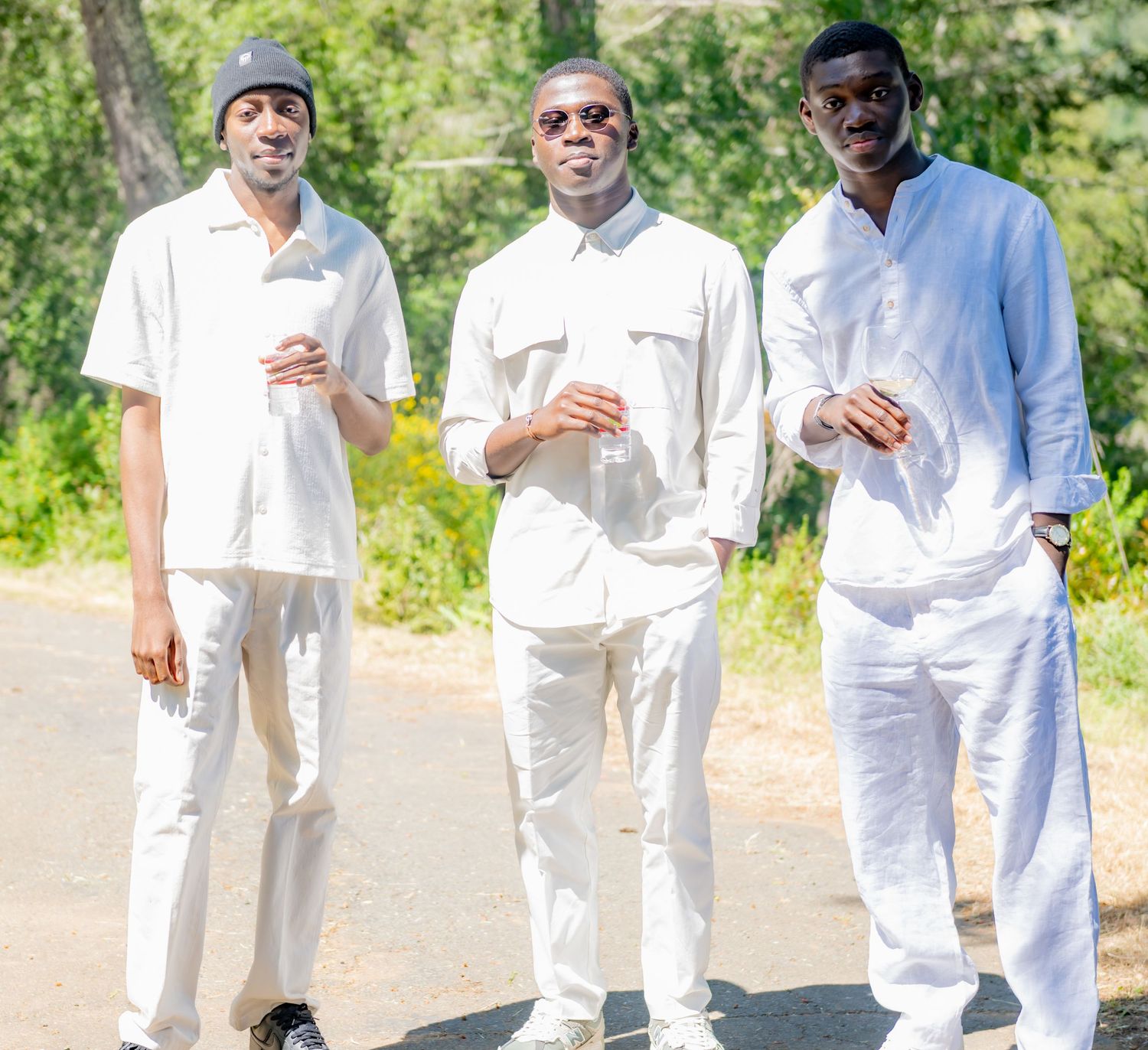 Three young men dressed in white outfits posing together on an outdoor path on a sunny day.