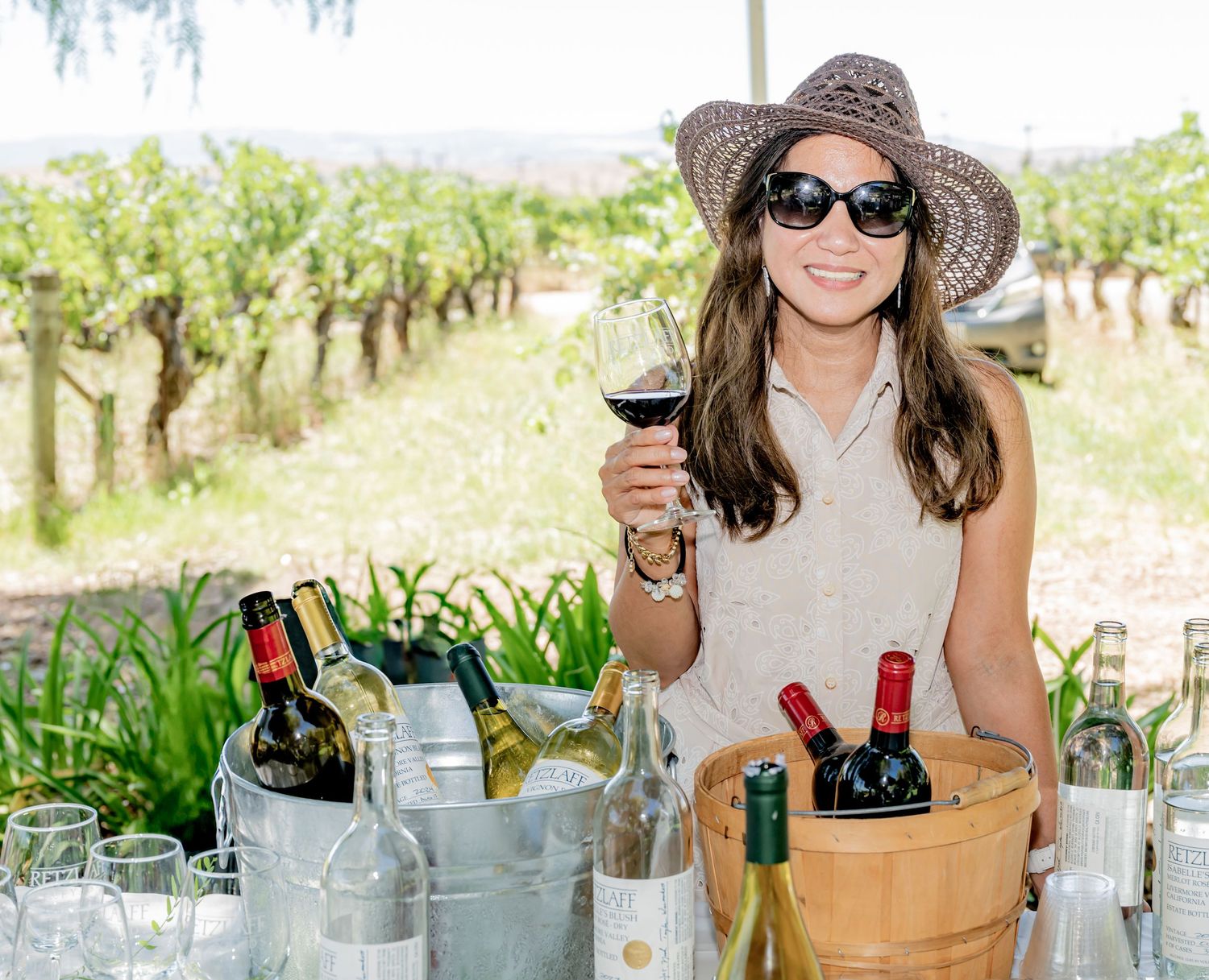Woman in sun hat holding red wine glass at vineyard surrounded by bottles in ice buckets on sunny day.