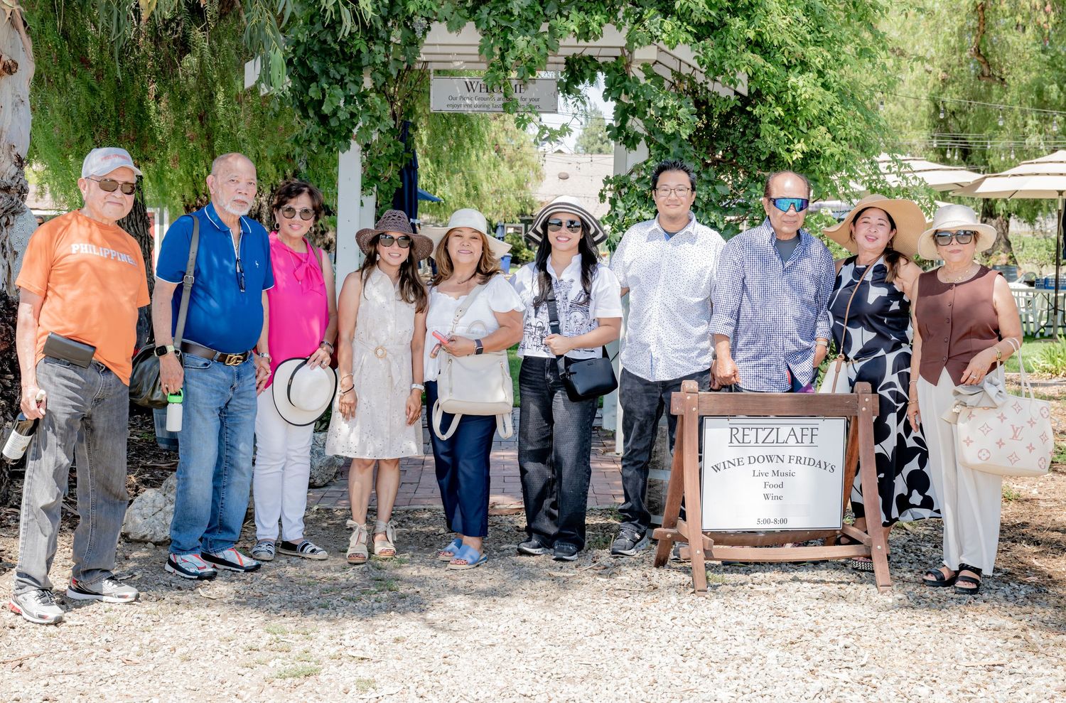 Group of ten people posing together outdoors near a rustic vineyard sign on a bright sunny day.