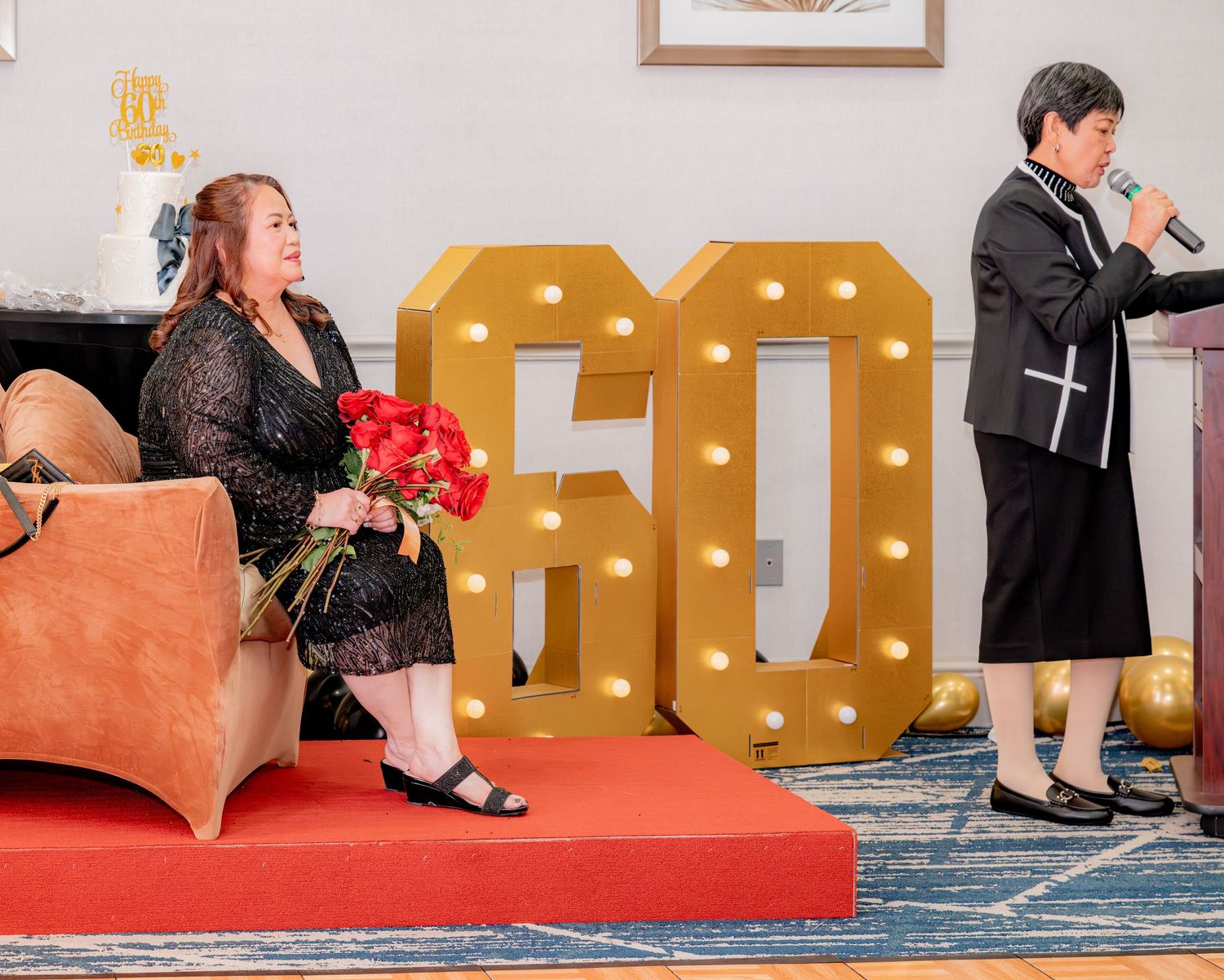 Woman seated with red roses beside illuminated 60 marquee letters at birthday celebration.