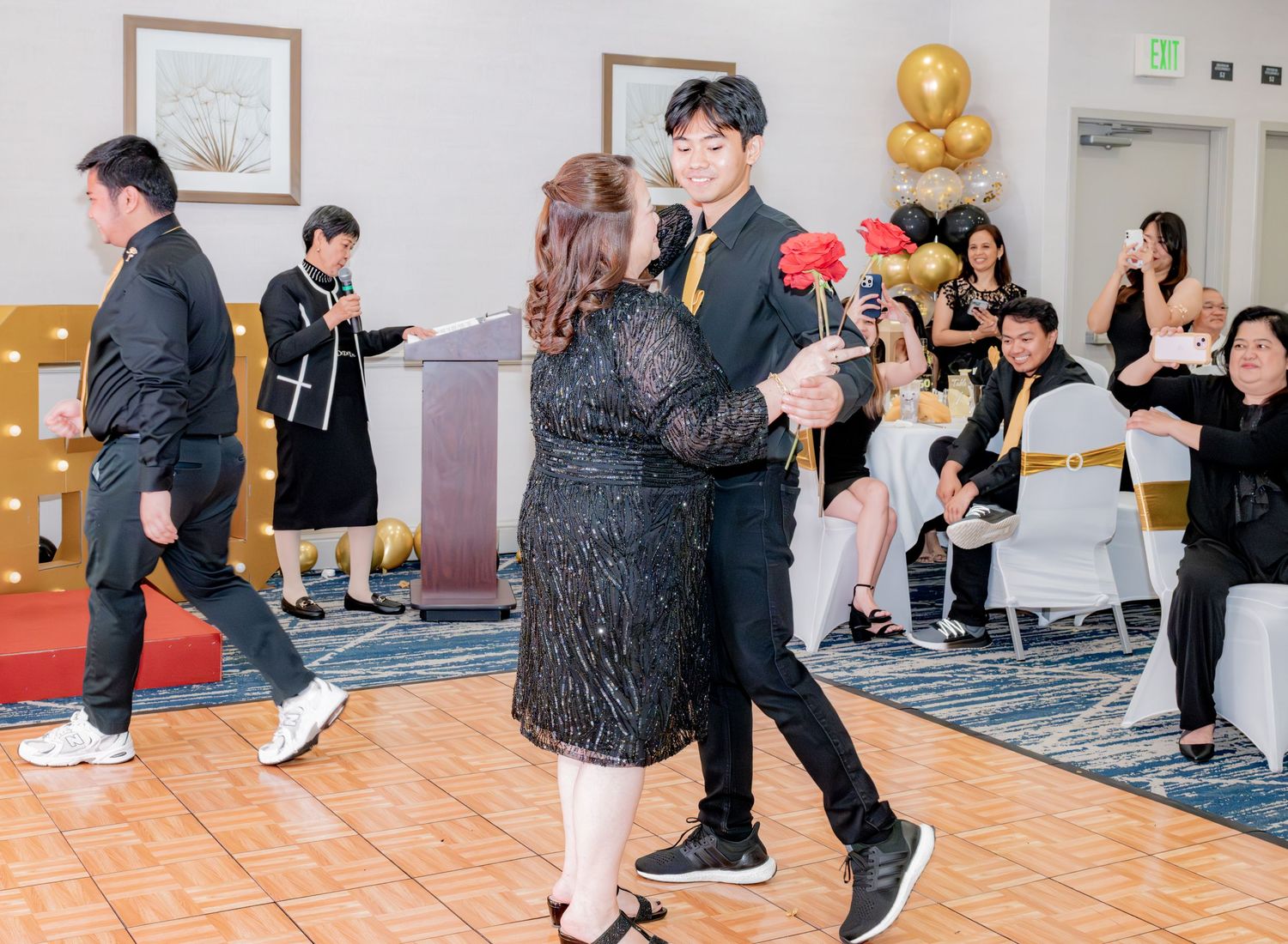 Young man in black suit dances with woman in black sequin dress at elegant 60th birthday party celebration.