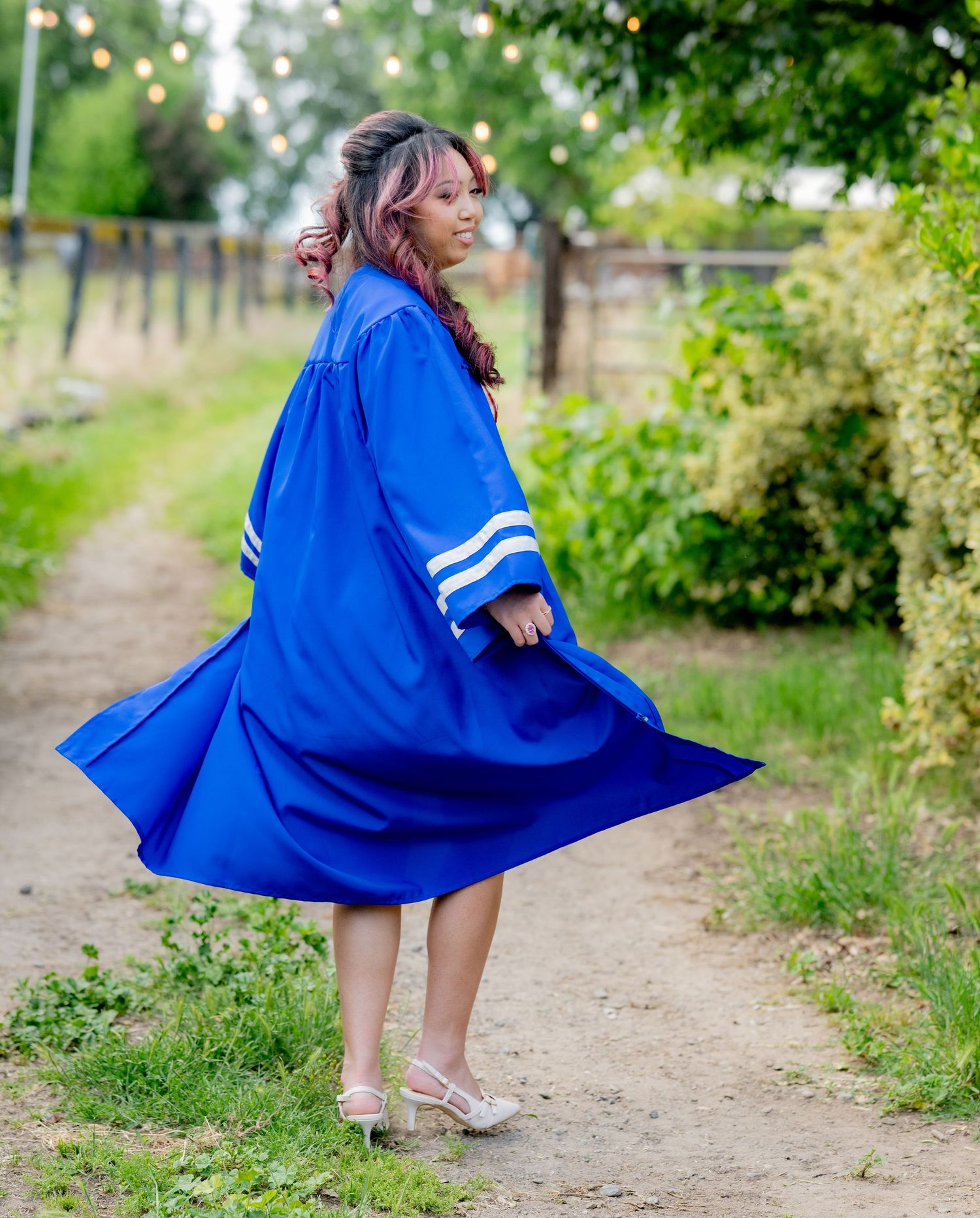 Graduate twirls her royal blue commencement gown on a rustic garden path surrounded by greenery.