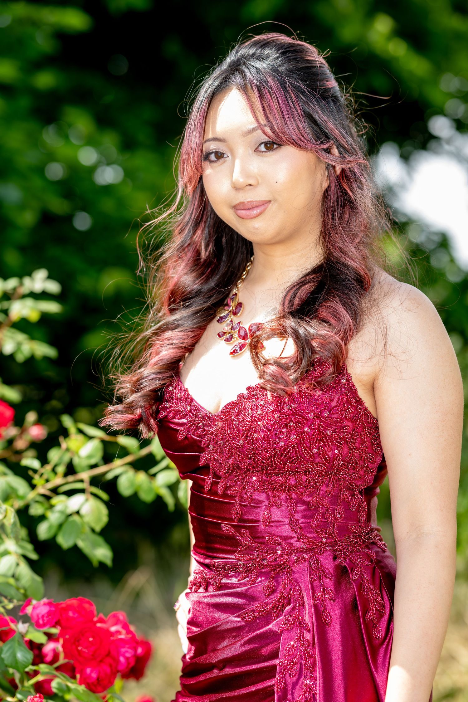 Woman in red lace satin gown poses among red roses in lush green garden.