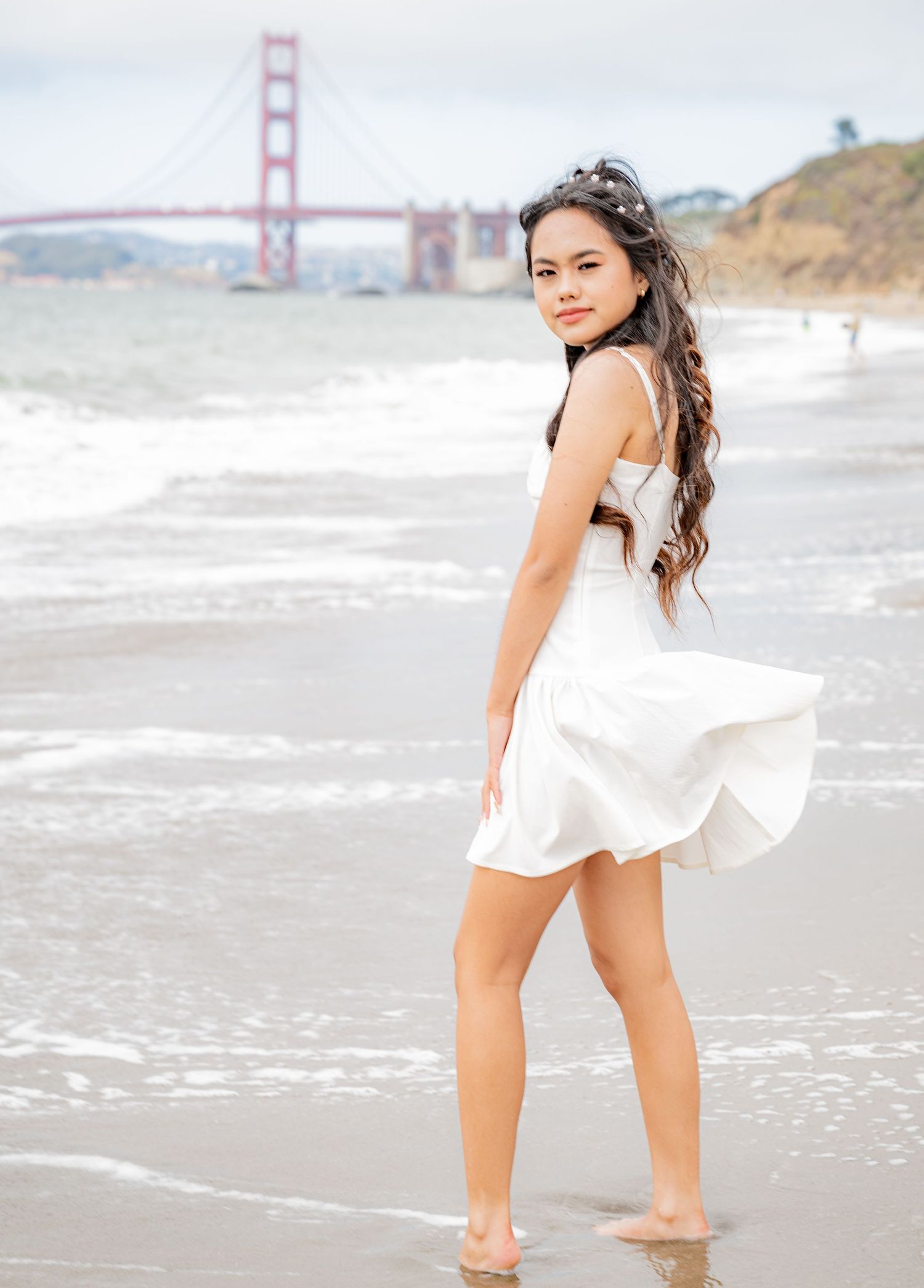 Young woman in white dress standing at Baker Beach with Golden Gate Bridge in background, waves nearby.