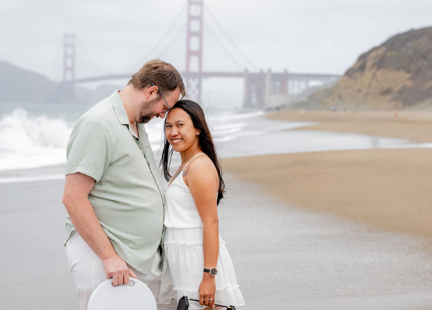 Couple sharing a tender moment on Baker Beach with Golden Gate Bridge visible in background.