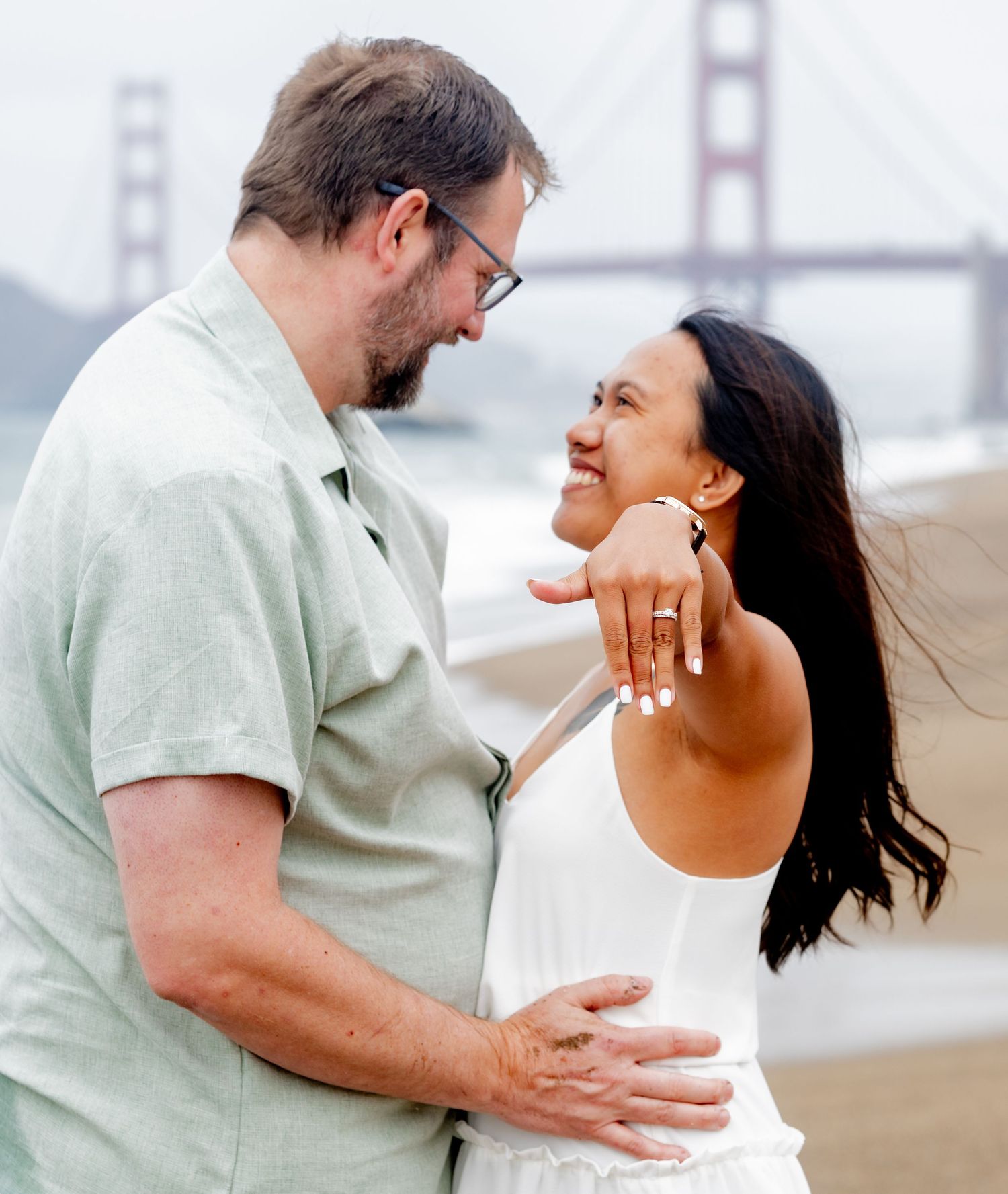 Couple smiling at each other on Baker Beach, woman showing engagement ring with Golden Gate Bridge behind.
