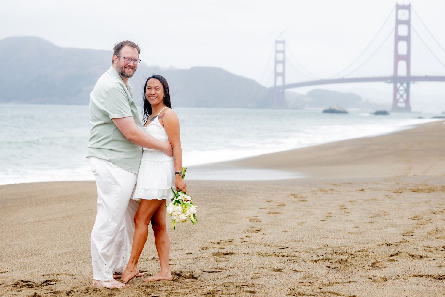 Couple posing on Baker Beach with Golden Gate Bridge backdrop, woman holding white bouquet.