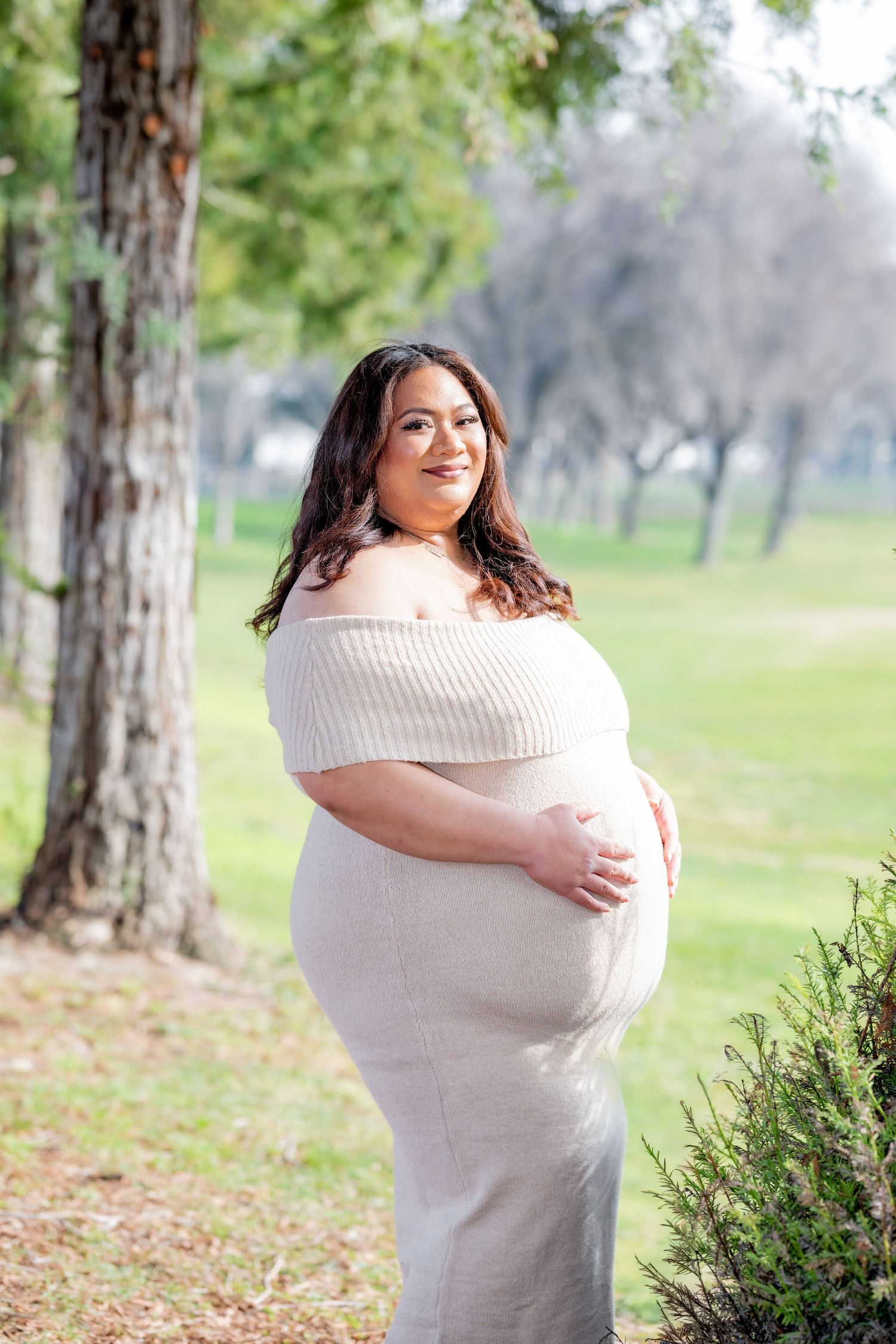 Glowing pregnant woman in off-shoulder cream knit dress smiles while cradling her baby bump in a park.