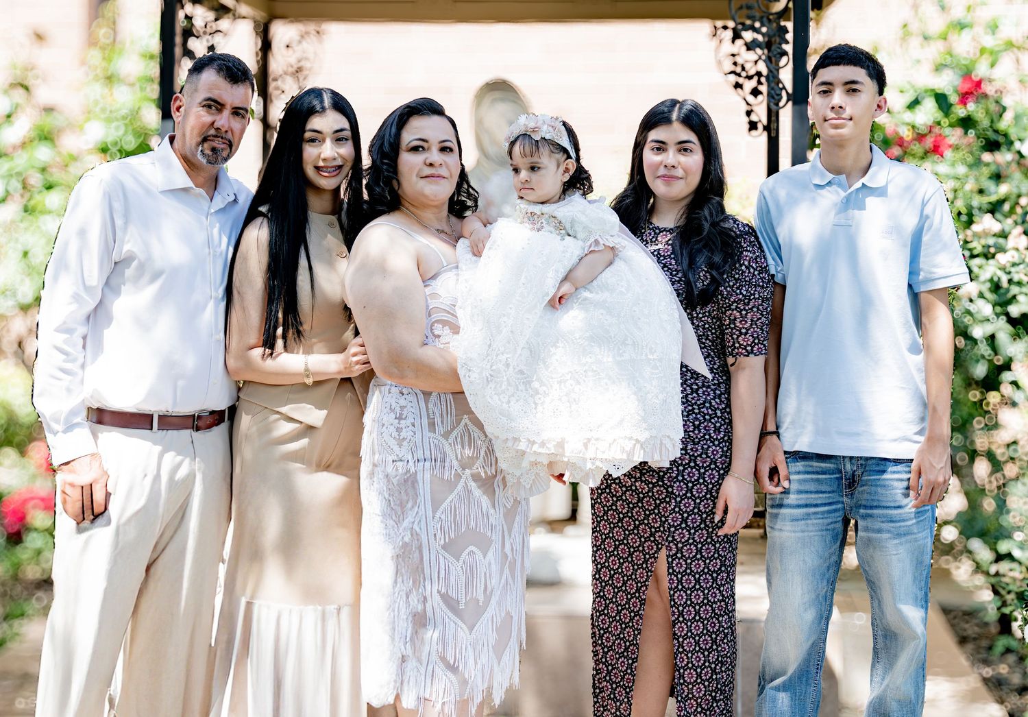 Extended family group portrait outdoors with baby in white baptism gown at garden setting.
