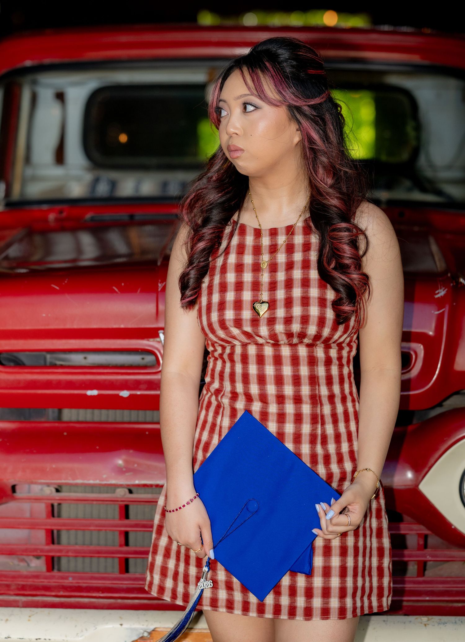 Young female graduate in red gingham dress holds blue mortarboard cap beside a vintage red truck.