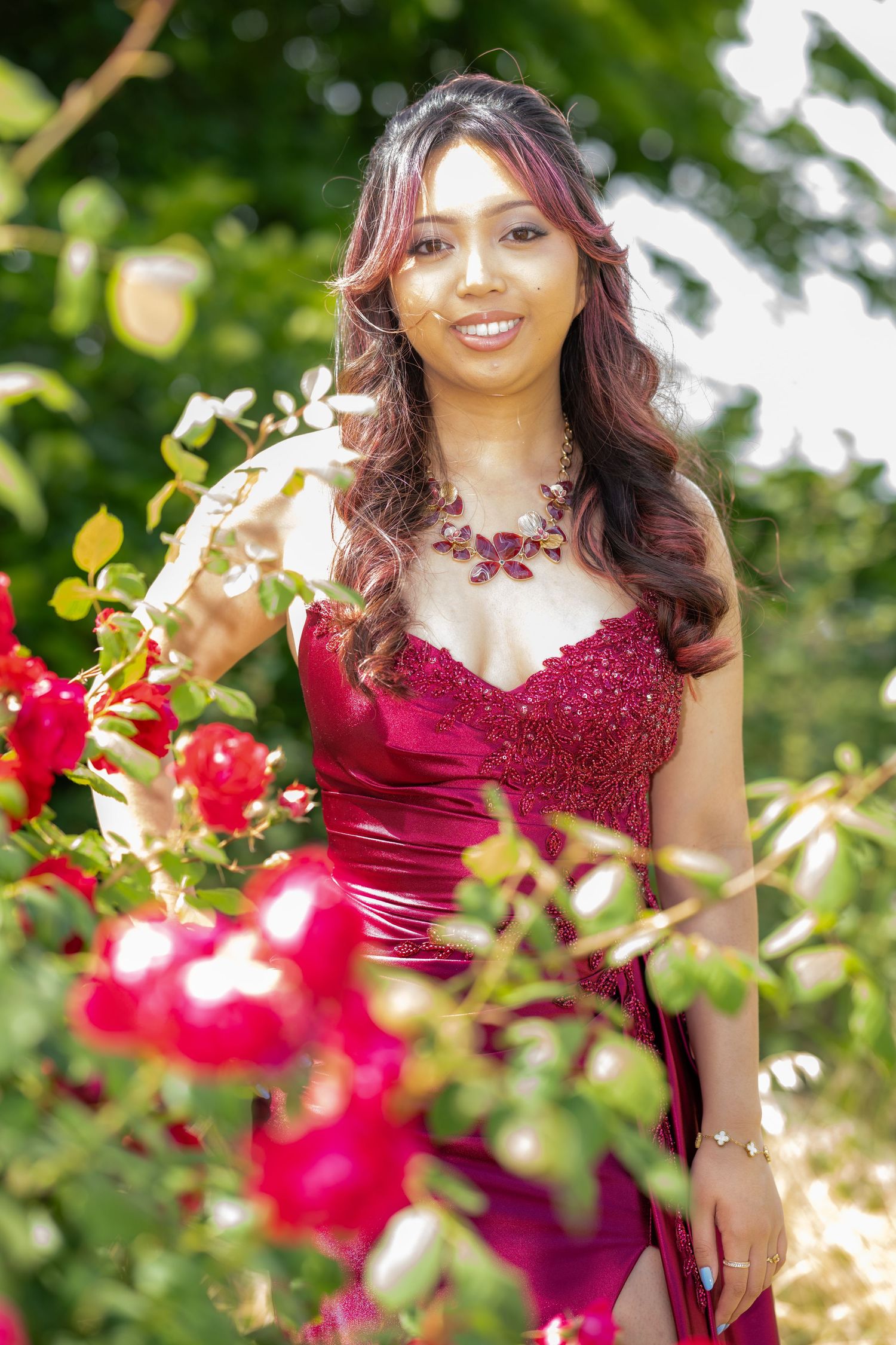 Smiling woman in red satin gown stands among blooming red roses in garden.
