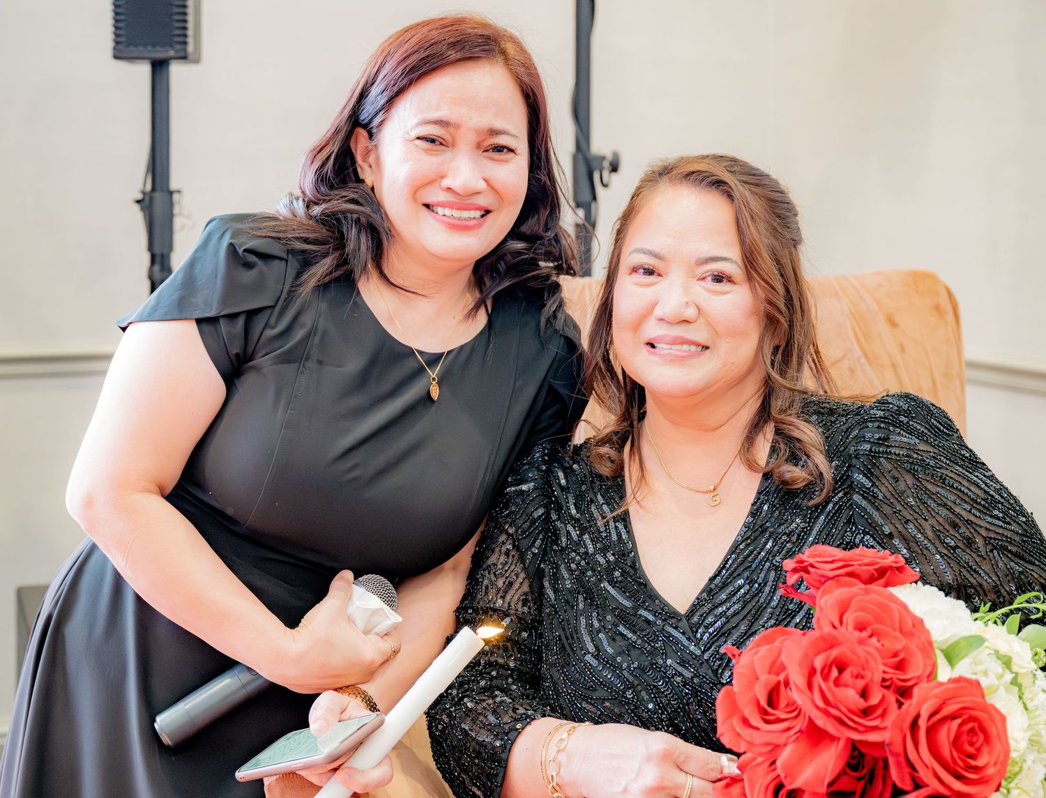 Two smiling women posing together at a celebration, one holding red roses and papers.