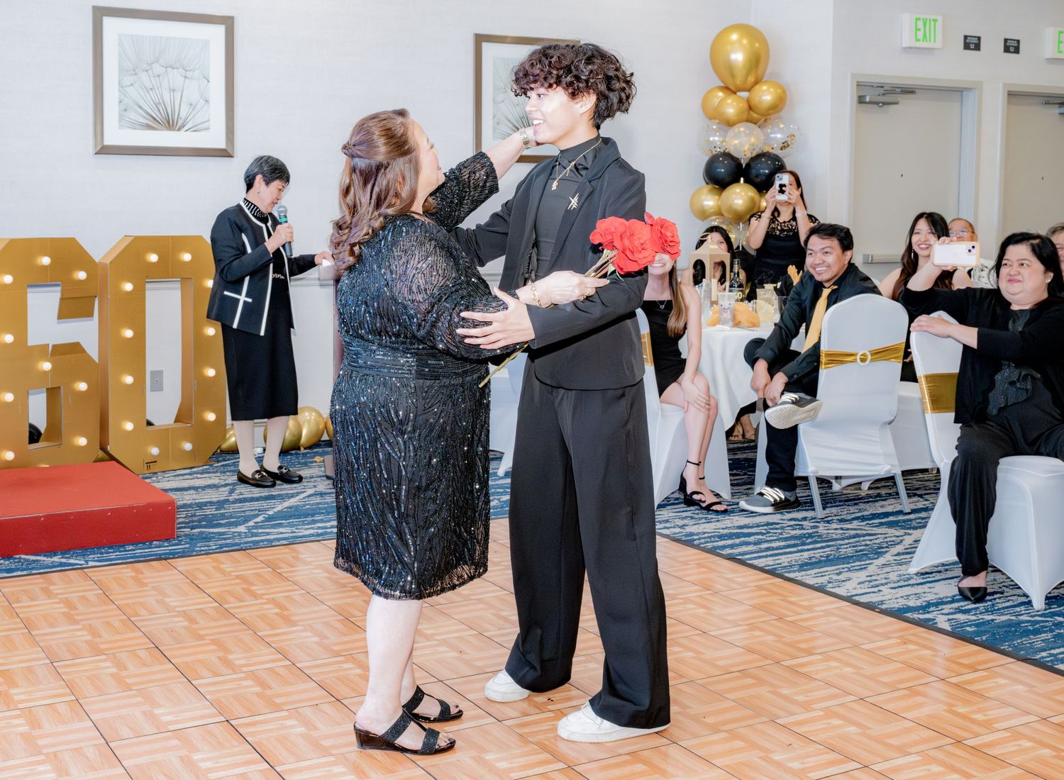 Young man in black outfit presents red roses to woman in sequin dress at 60th birthday celebration.