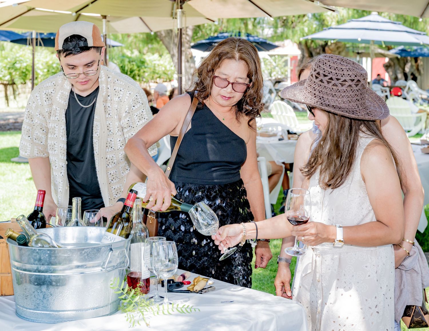 Woman pouring wine at an outdoor event with guests enjoying drinks under white umbrellas.