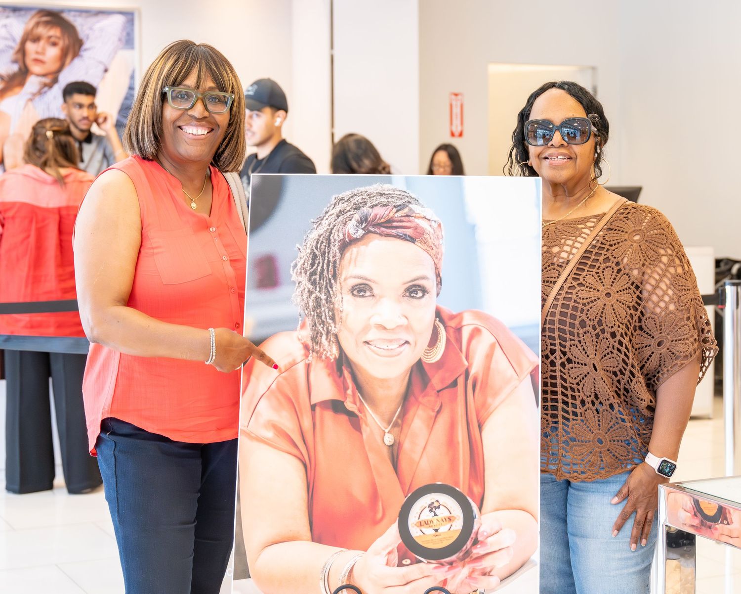 Two women posing with a portrait painting at an indoor art display event.