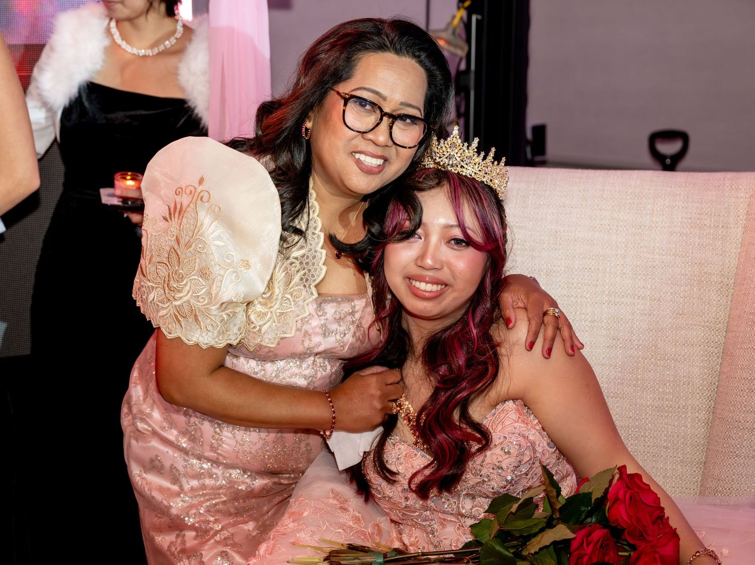 Two young women in pink formal dresses pose together, one wearing a tiara and holding red roses at a quinceañera.