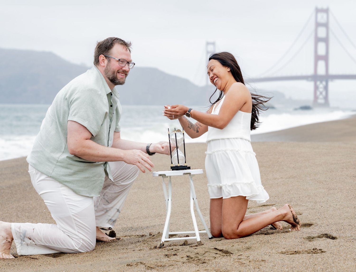 Joyful couple kneeling on Baker Beach near small white table, Golden Gate Bridge in background.