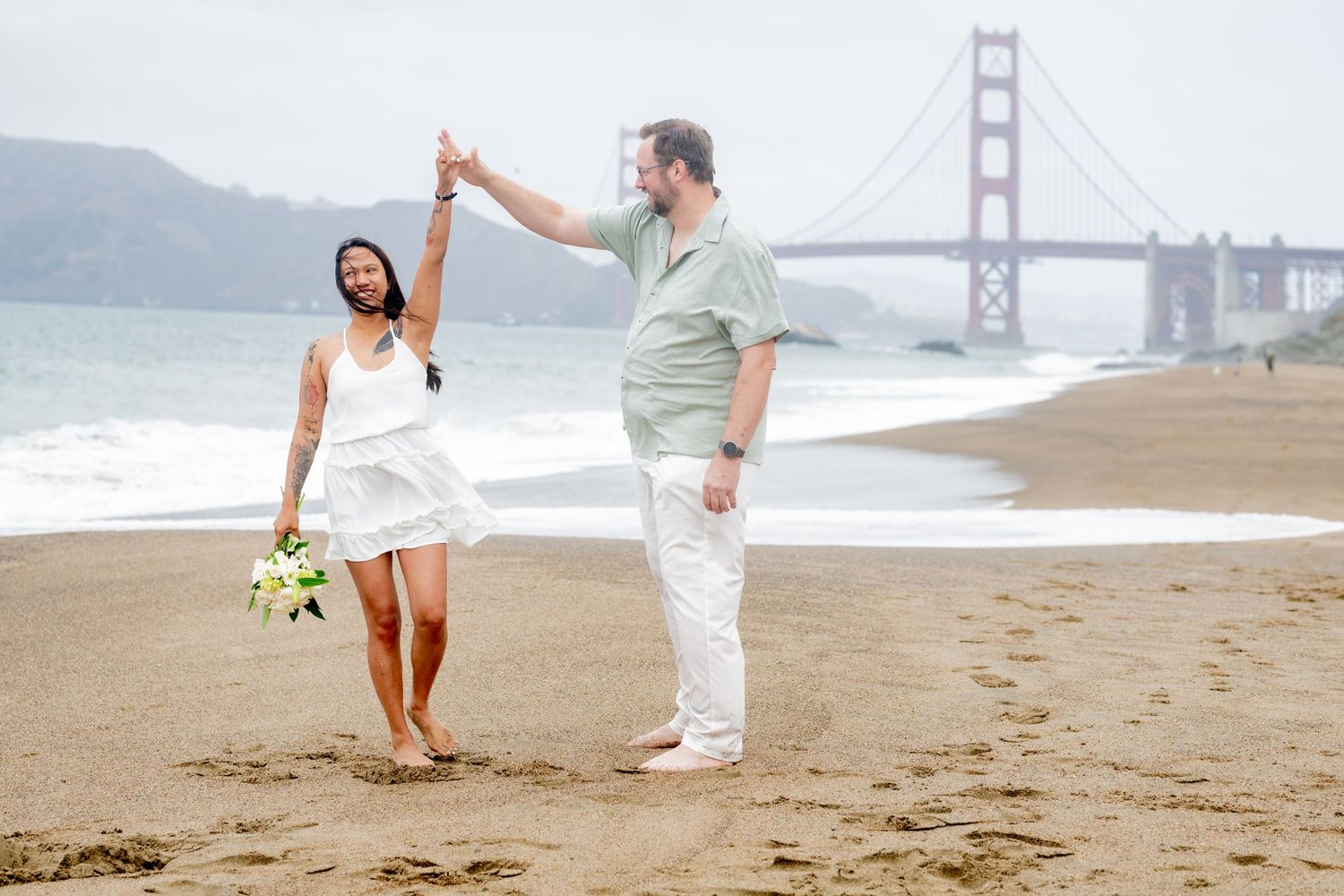 Man spinning woman on Baker Beach with Golden Gate Bridge visible through the misty background.