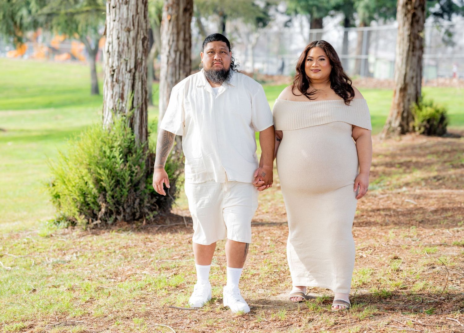 Expecting couple walks hand-in-hand through a park, woman in cream maternity dress and man in white outfit.