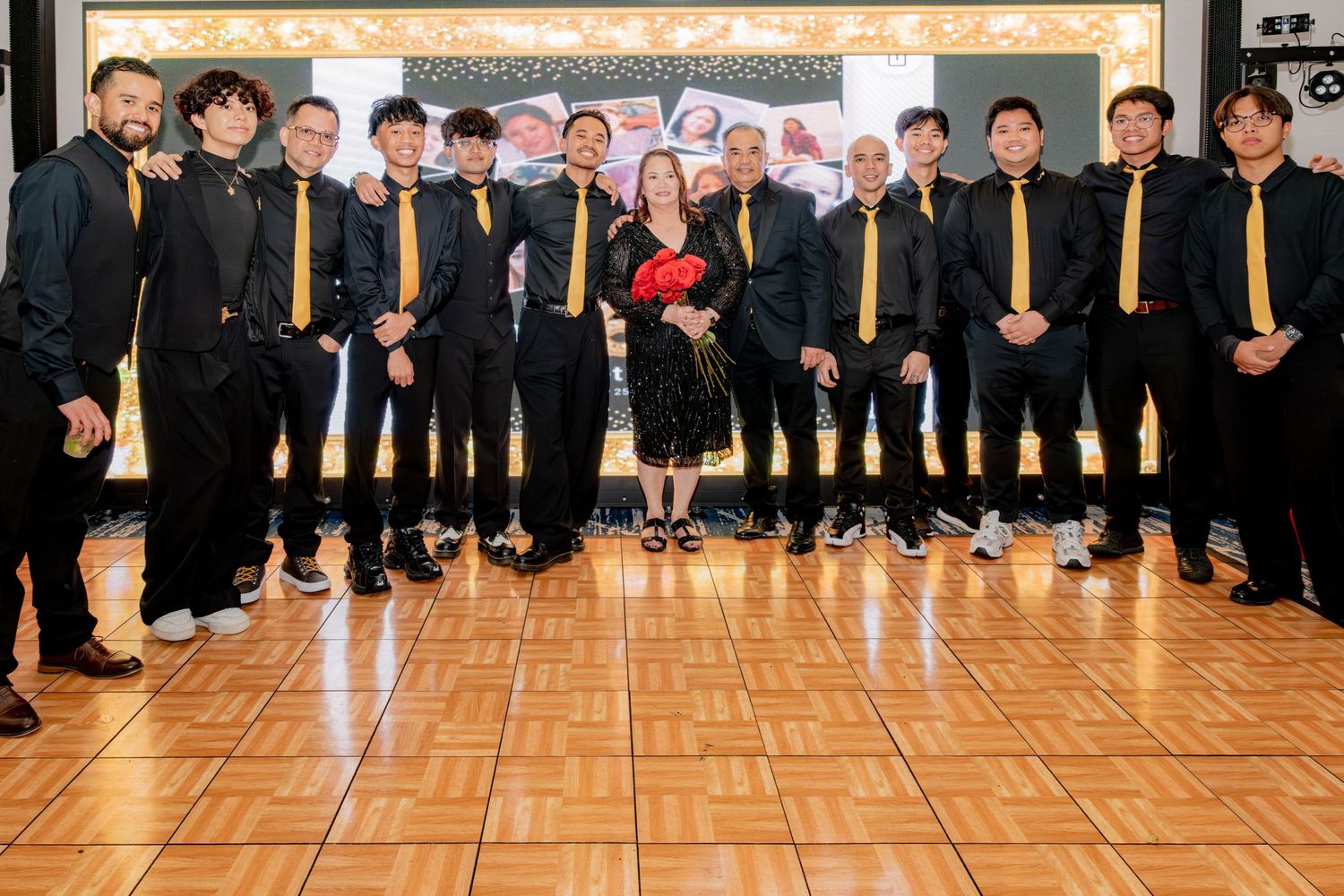 Group photo of men in black and gold attire with woman holding red roses at party.