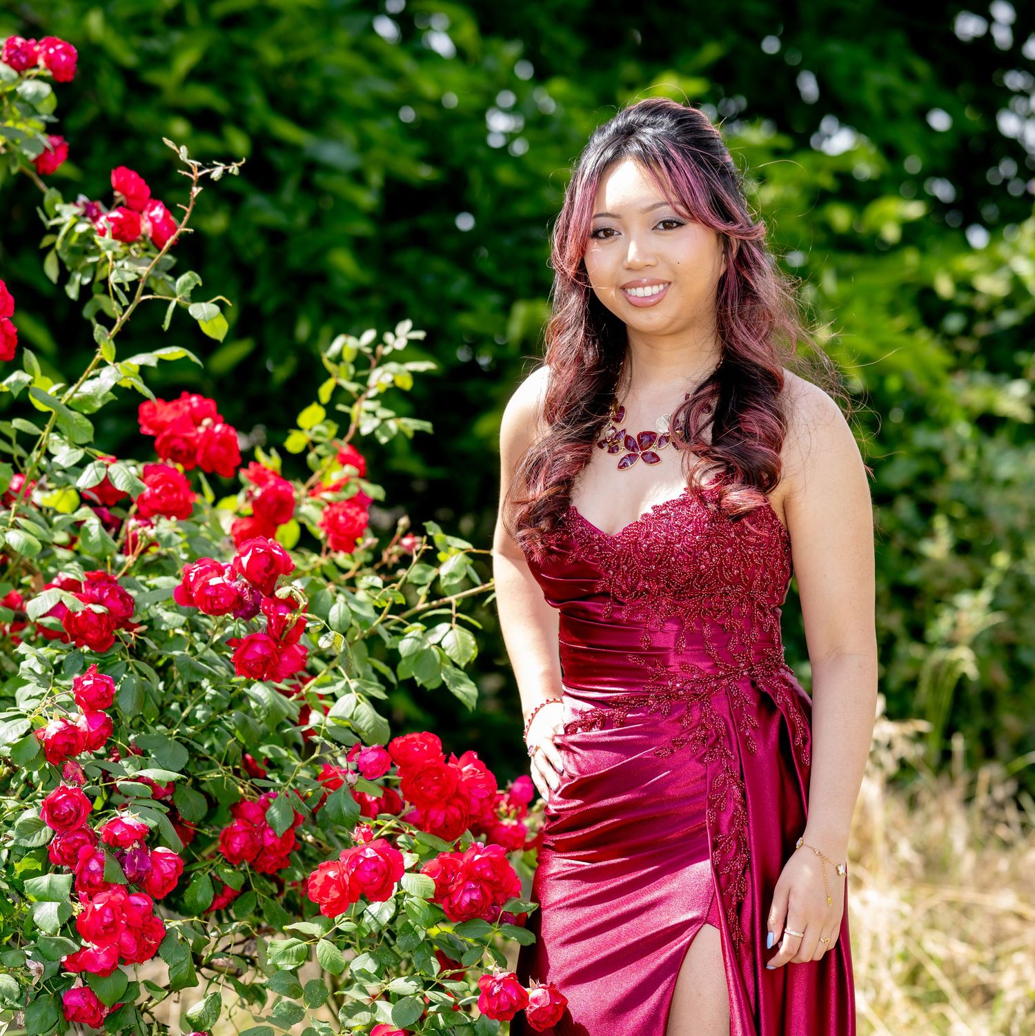 Young woman in red satin gown stands beside blooming red roses in garden.